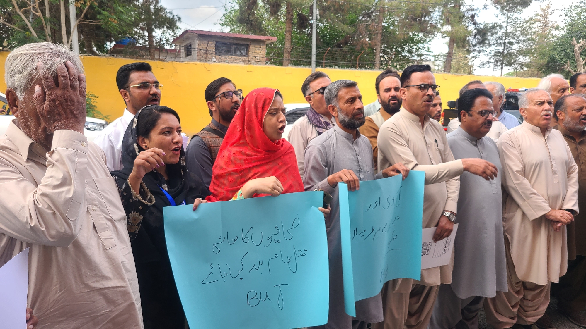 In Quetta City Tehsil, Pakistan, a group of individuals stands together outdoors, holding blue signs with Urdu writing, suggesting a demonstration or protest. The individuals are primarily middle-aged and older men, dressed in traditional South Asian clothing such as shalwar kameez and kurtas, in neutral tones like beige, gray, and white. There are also a few women, one of whom is wearing a red headscarf. One of the men covers his face with his hand, while others are speaking or shouting.
The background includes a yellow building, trees, and a cloudy sky. A few parked cars are partially visible. The overall impression is of a gathering expressing a shared concern, potentially political or social.
