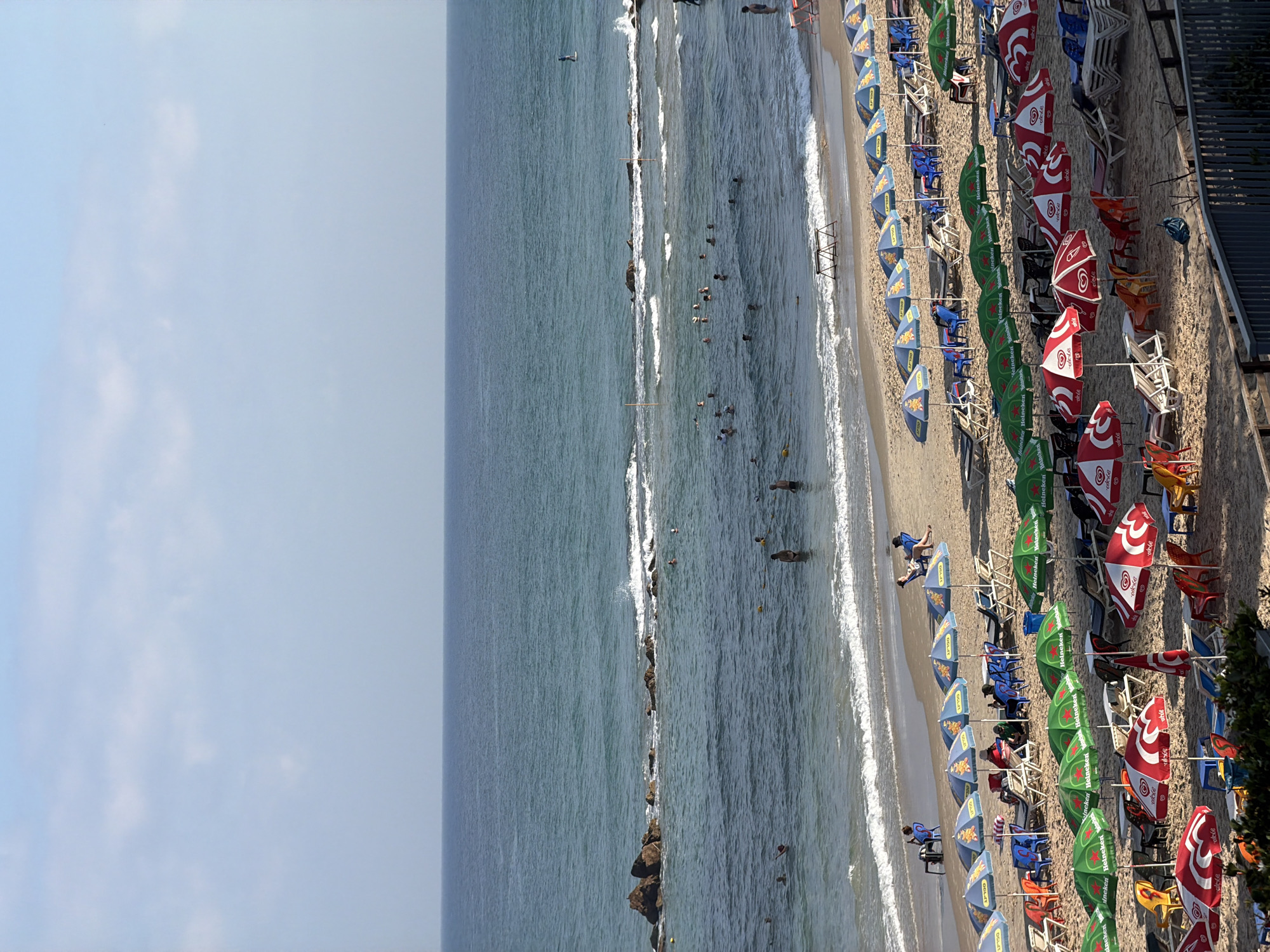 This image shows a crowded beach in Bat Yam, Israel, with many umbrellas and beach chairs set up on the sand. The ocean is visible in the background, with waves crashing on the shore. Several people are in the water, swimming and playing. Others are relaxing on the beach under the umbrellas. The beach appears to be a popular spot, with a lot of activity.