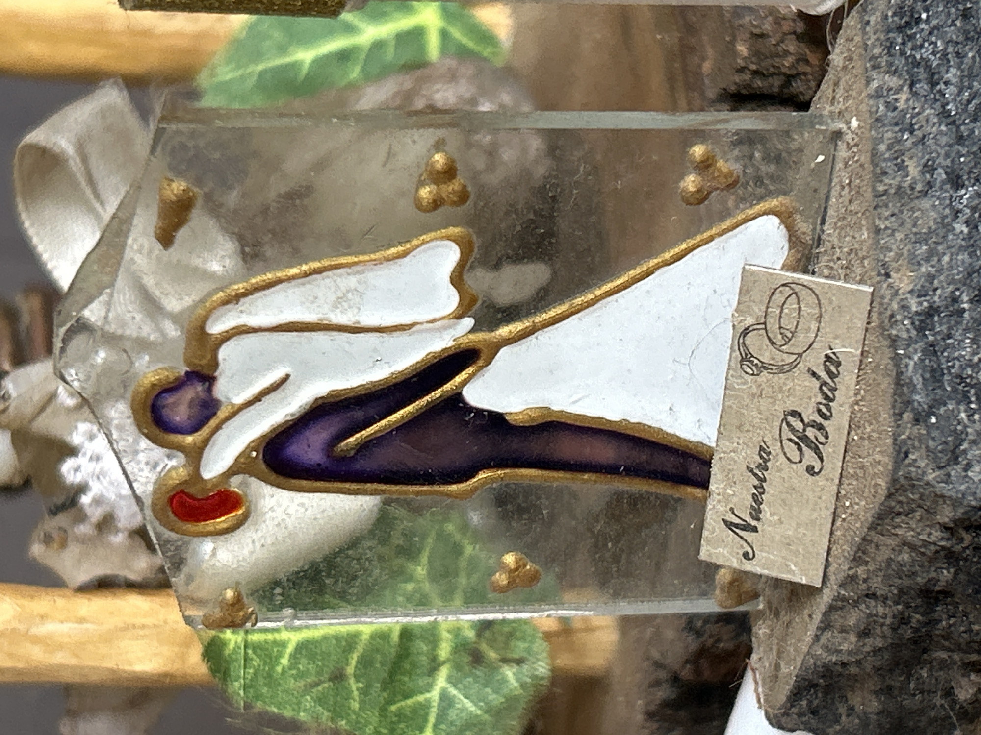 A close-up, eye-level shot captures a decorative figurine, likely a wedding favor, set against a blurred backdrop of rustic wood and greenery. The figurine, encased in clear material, depicts two figures in an embrace, rendered in purple and white with gold trim. The figures appear to be a bride and groom, with the bride wearing a flowing white gown. A small, tan-colored tag in the foreground reads "Nuestra Boda" and features a drawing of two intertwined wedding rings. The base of the figurine is mounted on a rough, wooden surface, possibly bark or a weathered piece of wood. The overall impression is that of a delicate, sentimental keepsake. There is no information available to indicate the location within the city of Santa Ana, Venezuela.