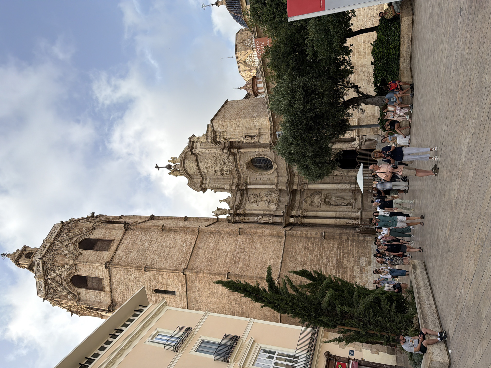 In the city of Unknown, a large, weathered stone bell tower, characteristic of Gothic architecture, dominates the left side of the frame, its intricate carvings and arched windows clearly visible against a sky with scattered clouds. To its left, a modern building with a pale facade and balconies with black railings stands, contrasting with the ancient tower. In front of the tower and a portion of a stone church is a wide, paved plaza, bustling with activity. A diverse group of people are walking in various directions across the plaza, some in small groups, others alone.
Several individuals are seated on a low stone wall in the mid-ground, under the shade of trees. One man in a gray t-shirt and black shorts is sitting alone on the edge of a planter, looking at his phone. A couple, a man in a light pink shirt and shorts and a woman in a blue and white striped outfit holding cameras, are standing and talking in the foreground. Another group of people are seated on a bench in the background. Behind the people and trees, the stone facade of the church is visible, featuring elaborate sculptures and an arched entrance. A red banner with white text is partially visible on the far right. The overall scene captures a moment of everyday life in a historic urban setting, with tourists and locals interacting amidst impressive architecture.