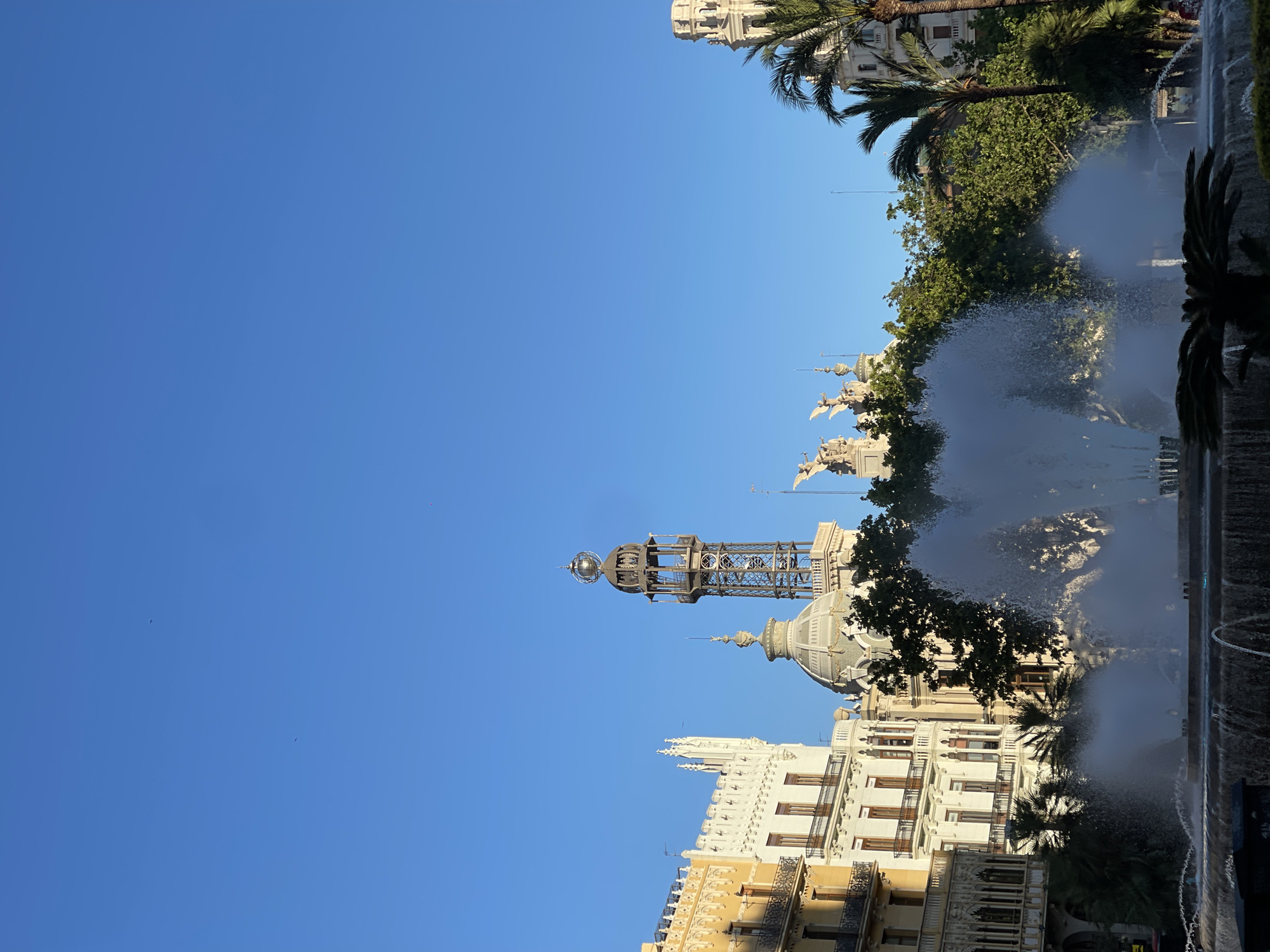 The image is a ground-level shot from the Plaza del Ayuntamiento in Valencia, Spain. The Post Office building (Edificio de Correos), as well as fountain, are visible. The blue sky dominates the upper portion of the image. Fountain spray partially obscures the view of the architecture. Palm trees and other green foliage are scattered around the periphery. The image appears to have been taken in the daytime, under clear weather conditions.