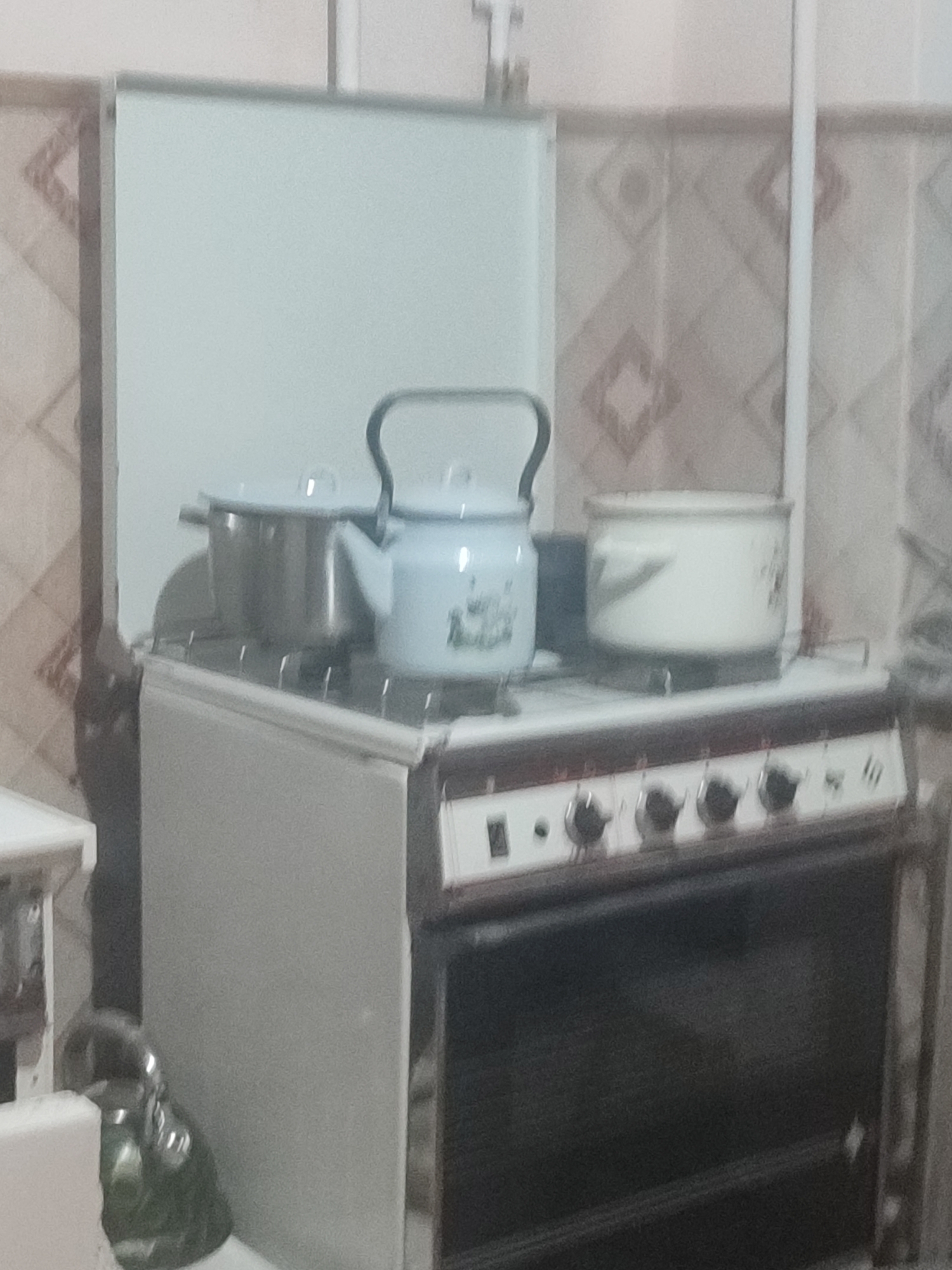 An interior scene depicts a domestic kitchen setting. In the foreground, a white gas stove with a black oven door is visible. The stovetop features a metal grate, on which two pots and a light blue enamel kettle are placed. The left pot is made of stainless steel, while the pot to the right is white with a subtle pattern. The kettle has a black handle and a spout. To the left of the stove, a portion of a white countertop or cabinet is visible, with a dark green metal object, possibly a smaller kettle, on it. The background shows a tiled wall with a diamond pattern in beige and brown. The lighting in the room appears to be artificial. The image is slightly blurry. No people are visible. The location of this scene is unknown, but it is a general depiction of a kitchen in Chernivtsi, Ukraine.