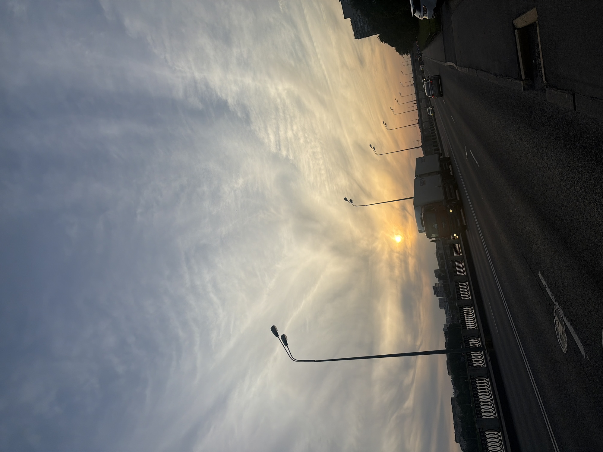 The image captures a wide-angle outdoor scene on a multi-lane bridge or highway during either sunrise or sunset, indicated by a low-lying orange sun near the horizon and warm hues in the lower sky, contrasting with cooler blue-grey tones and wispy, streaky cirrus clouds higher up. The foreground elements are heavily silhouetted due to backlighting.
In the mid-ground, a paved, multi-lane asphalt road extends from the bottom of the frame, marked with white dashed and solid lines. A large white box truck with an orange cab is visible on the left lane, its headlights illuminated, moving away from the viewer. Further down the road, a silver sedan and other indistinct vehicles are also traveling away. On the far right, a partial view of a white SUV is seen parked or stopped on a paved sidewalk.
Numerous tall, dark streetlights with dual, upward-curving fixtures line both sides of the road, extending into the distance. On the left side of the road, a decorative metal railing with a repeating oval pattern separates the roadway from a broad body of water, likely a river, which reflects the sky. Across the water, a cityscape consisting of silhouetted multi-story buildings and trees is visible on the distant bank. On the immediate right, a pedestrian walkway/sidewalk features a drain grate and is bordered by dark trees and foliage, also silhouetted. A circular manhole cover is visible on the road in the lower-left quadrant. The scene depicts typical urban infrastructure in an unknown city within Russia.