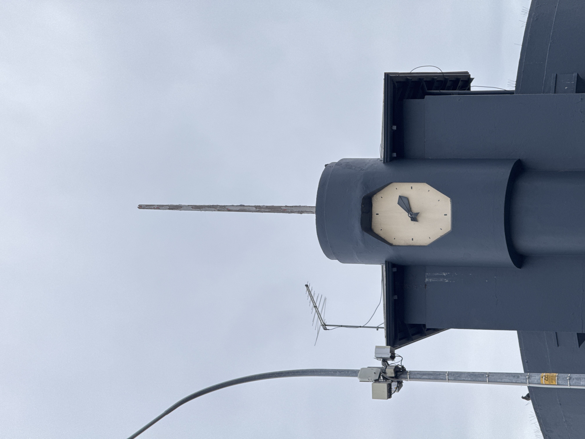 The image captures a close-up of a dark grey, octagonal clock face mounted on a cylindrical structure. The clock has cream-colored numerals and black hands, with the time appearing to be approximately 4:45. The structure is topped by a thin, wooden pole with peeling paint. To the left of the clock tower, a grey metal pole with attached electrical boxes and an antenna is visible. The background consists of a pale, overcast sky. A bird is perched on the lower section of the grey pole, and another small dark bird is on a ledge in the lower right of the frame. The scene is suggestive of a coastal or industrial area in Perth, Australia, likely near a public space or building with a prominent clock.