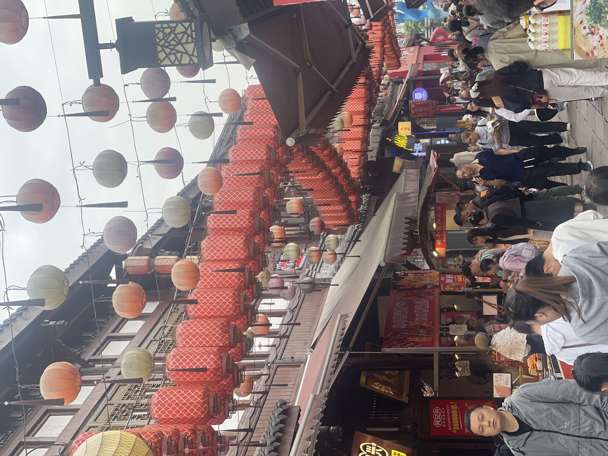 The image shows a street scene in Shapingba District, China. Red and orange lanterns are strung above the street. The buildings are constructed in a traditional Chinese style. People walk through the crowded street. There are a variety of shops and restaurants. Some of the signs are in Chinese, indicating the local food.