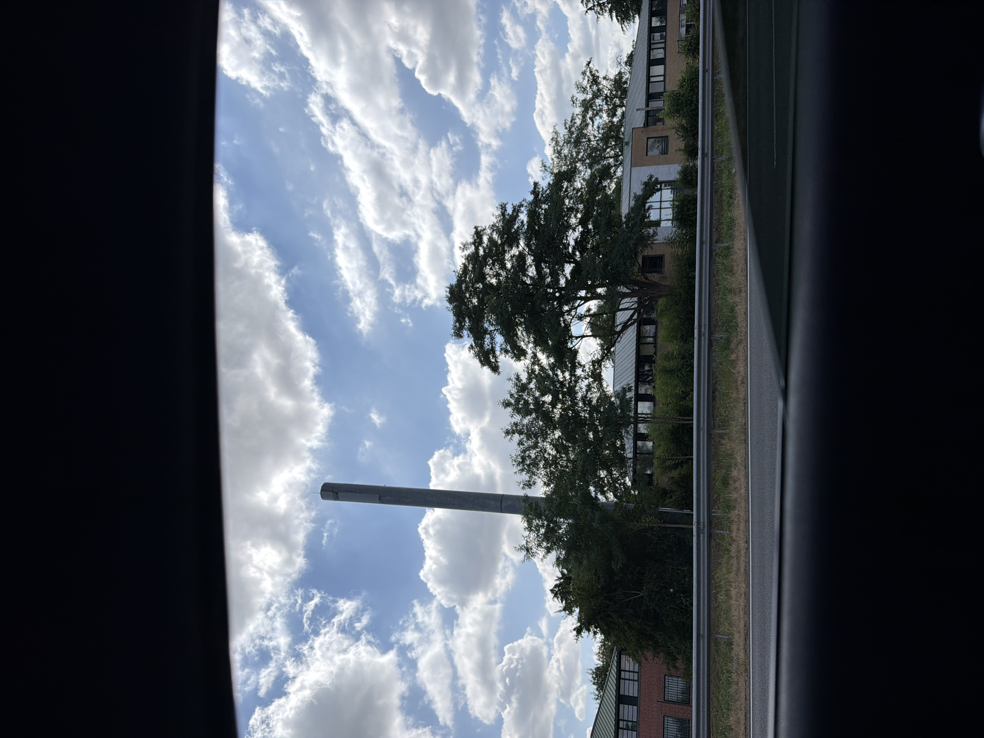 The image, taken from inside a vehicle, captures a view of a modern building complex with a distinctive tall, slender smokestack or ventilation tower in the foreground. The building appears to be a school or office building, with a long, low-rise structure featuring numerous windows. Mature trees are abundant around the complex, some partially obscuring the facade of the building. A metal guardrail separates the building area from what appears to be a road or highway in the foreground. The sky above is a bright blue with fluffy white clouds, suggesting a clear, possibly sunny day. The perspective suggests the photo was taken while driving past the location. This scene is consistent with the vicinity of a large institution or industrial area within a suburban or semi-industrial part of Évry-Courcouronnes, France, possibly near a transport artery.