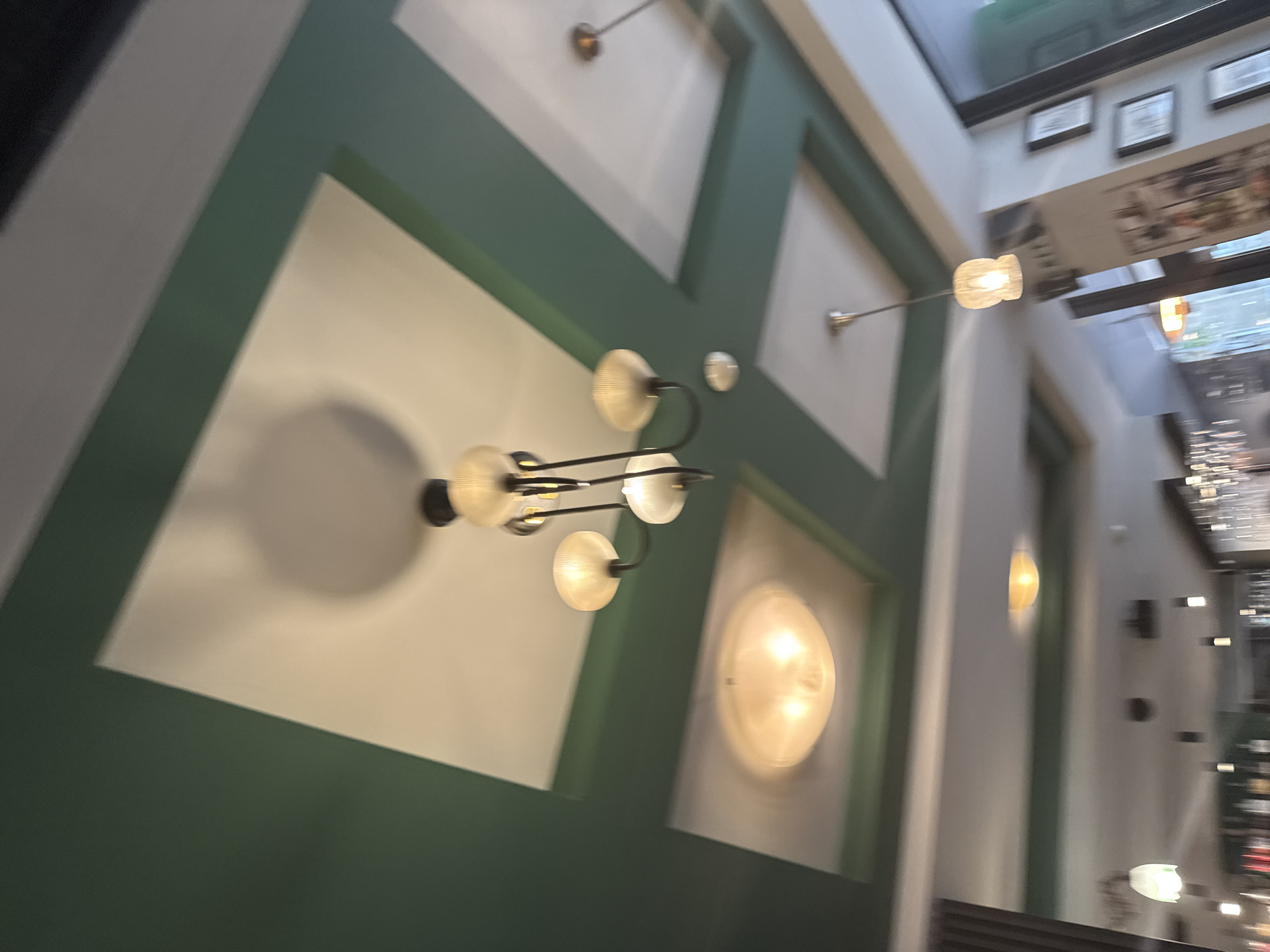 The image shows the interior of a building, likely a restaurant or bar, featuring distinctive wall decor with white rectangular panels bordered by green trim. A chandelier with multiple frosted glass orbs hangs from the ceiling. Other lighting fixtures, including wall sconces, are visible, suggesting a deliberately designed and illuminated space. Through an opening in the ceiling, there is a partial view of the exterior, which appears to be an urban setting. Signs and framed pictures are also visible. Due to the limited detail, it is not possible to identify the exact location in Sofia, Bulgaria, without further information.