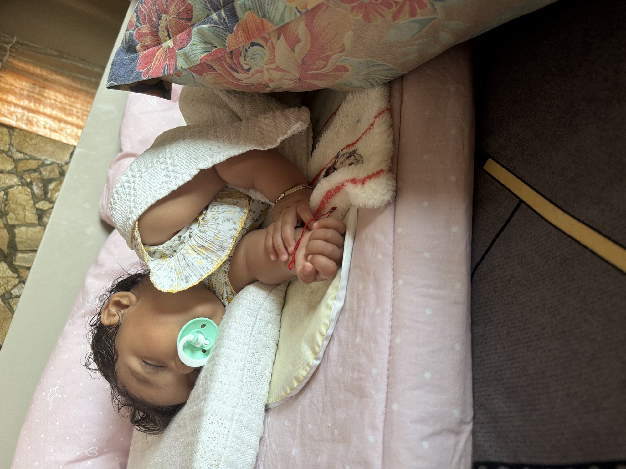 A baby is asleep on a pink polka dot patterned blanket in Razhevo Konare, Bulgaria. The baby is lying on its right side with a pacifier in its mouth and is covered by a white knitted blanket. The baby is wearing a yellow and white floral patterned dress. There is a pillow under the baby's head with a pink cover. Next to the baby is a colorful floral patterned cushion. The background shows a stone wall and curtains.