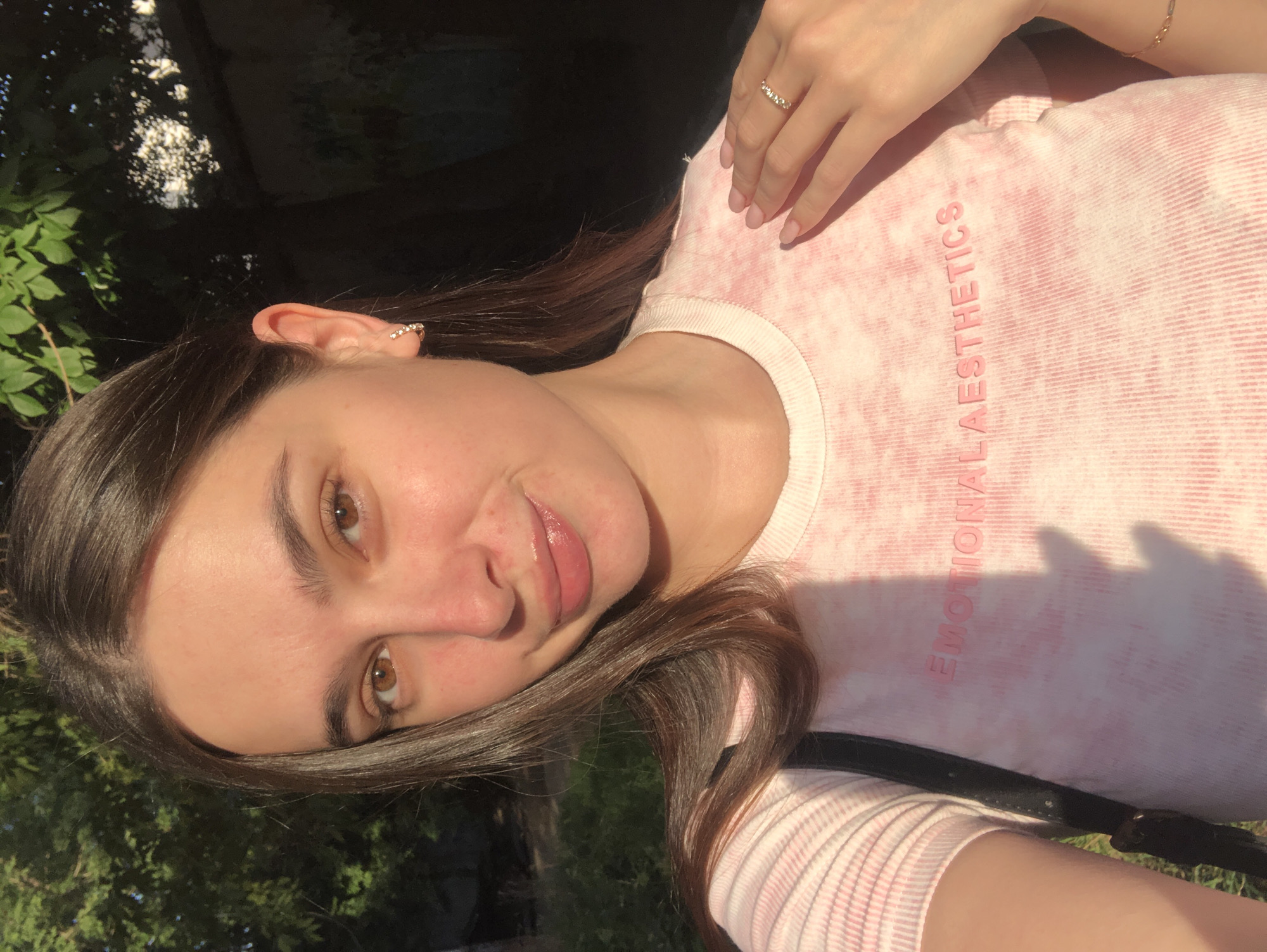 A young woman with long, dark brown hair, brown eyes, and light skin is taking a selfie outdoors during the daytime. She is wearing a light pink, ribbed t-shirt with the words "EMOTIONAL AESTHETICS" printed in a darker pink color. She has a small, diamond hoop earring in her left ear and a ring on her right hand, which is resting on her chest. The background appears to be a park or green space with trees and dappled sunlight. The image does not contain any information about the location in the city of Chaikovsky, Russia.