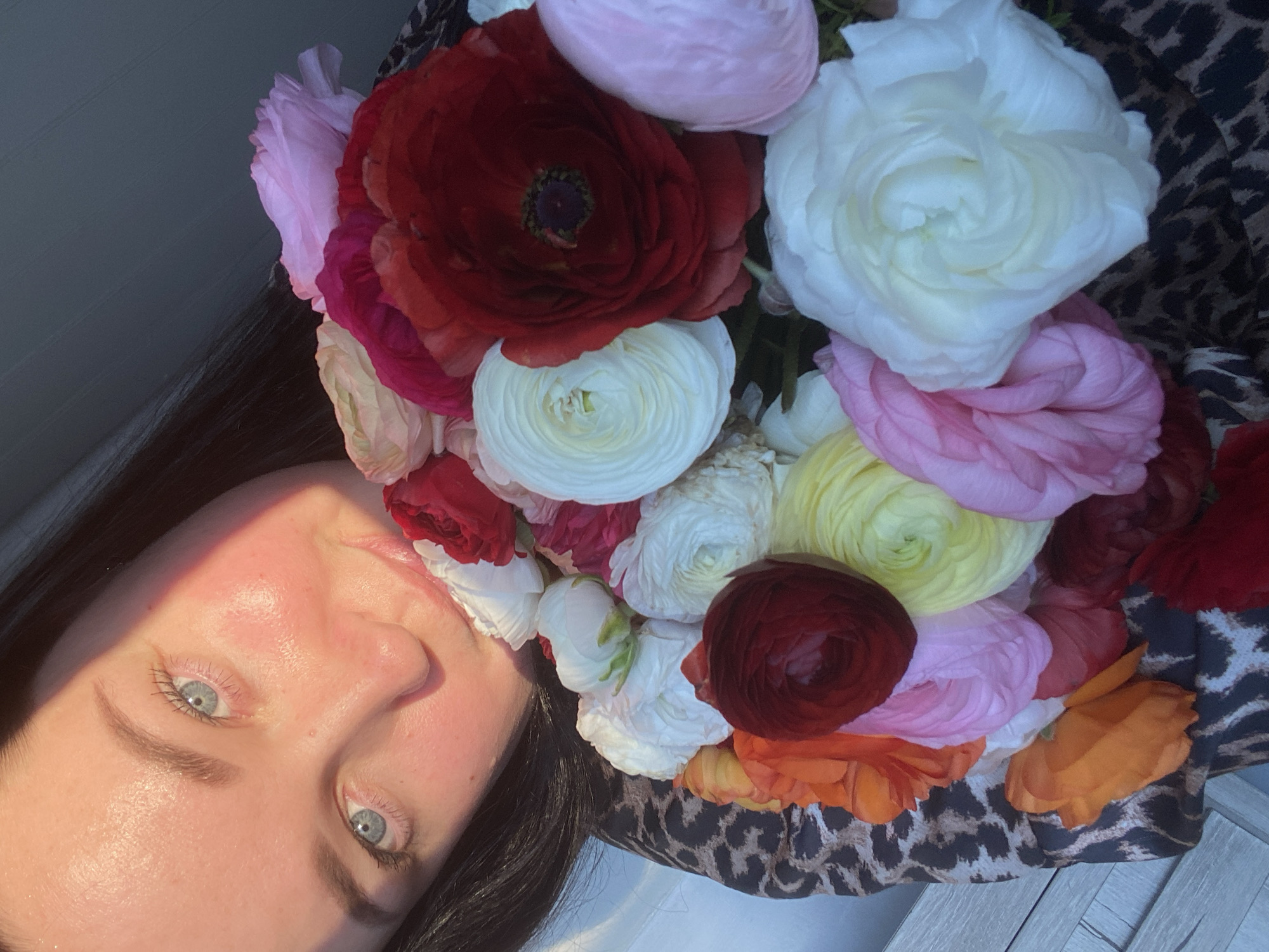 The image shows a woman's face, slightly angled, with a large bouquet of colorful ranunculus flowers positioned above her. The flowers include shades of white, pink, red, yellow, and orange. She has fair skin, light blue eyes, and dark hair. Part of her shoulder is visible, covered with an animal-print fabric. The lighting is bright, with a direct sunbeam across her face. The location, Sochi, Russia, cannot be confirmed or denied based on the image alone.