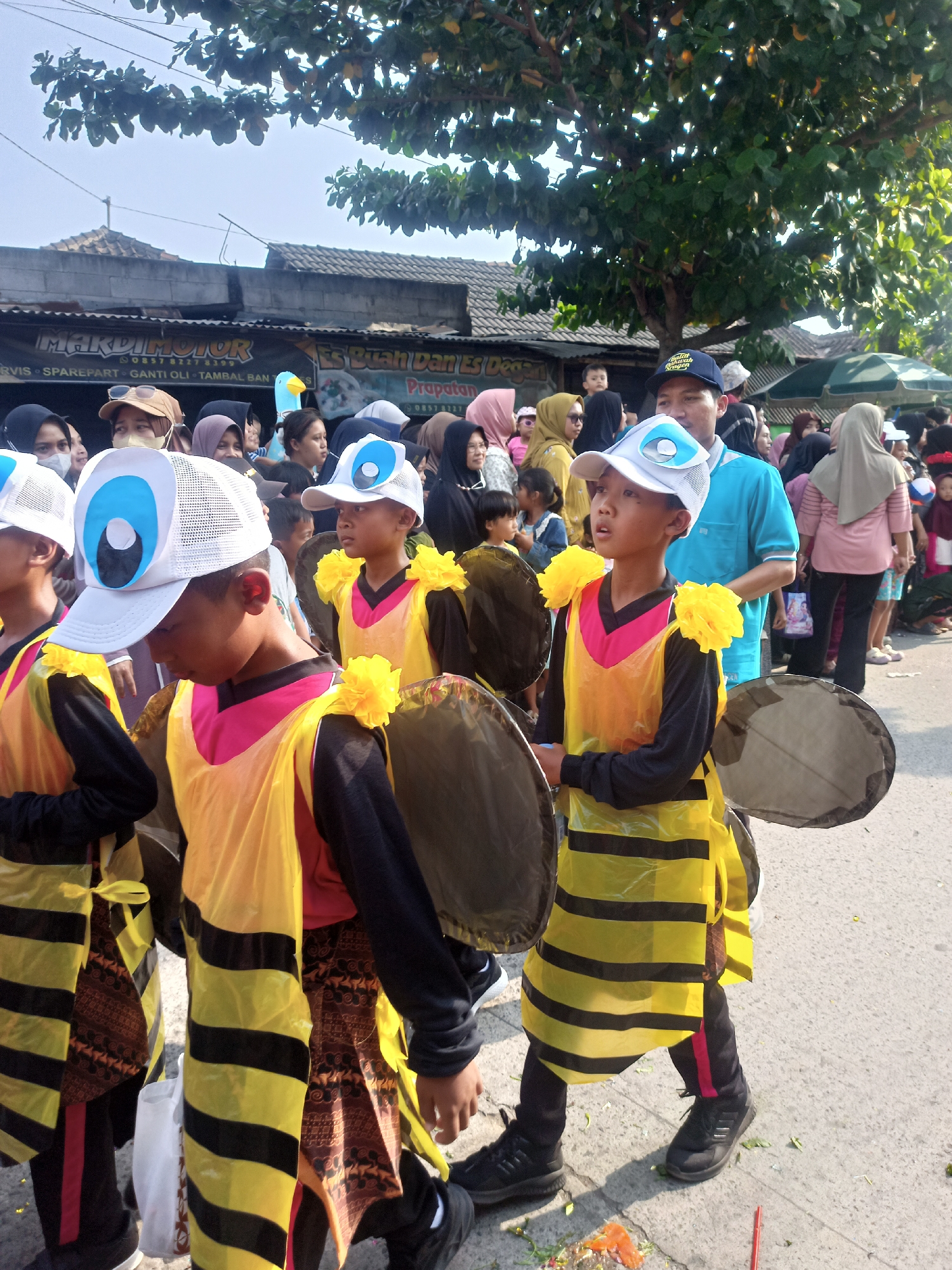 In Saren, Indonesia, a procession is underway. Several children are dressed in bee costumes, complete with yellow and black striped tunics and attached black wings. They are wearing white caps with eye designs. Some adults and other children are observing the procession, many wearing headscarves. In the background, there are buildings, trees, and awnings, indicating an outdoor urban or semi-urban setting. The ground is paved and shows some scattered debris, possibly from the event. The overall atmosphere appears festive.