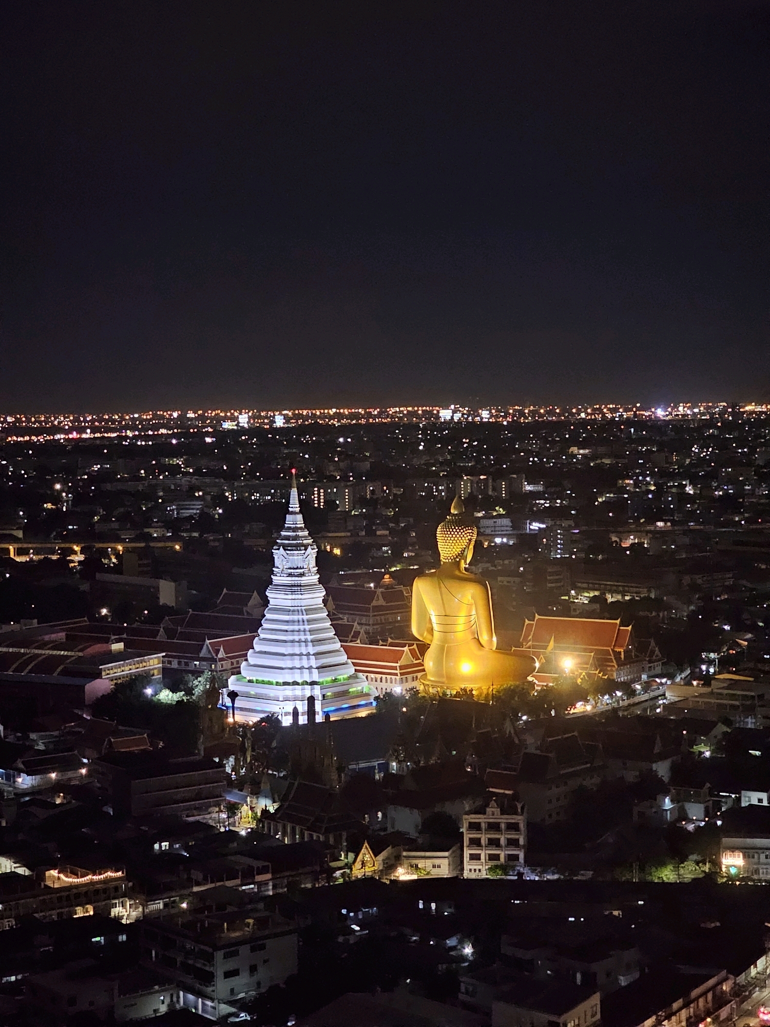 The image is a night-time, high-angle aerial shot of a cityscape in Bangkok, Thailand. Dominating the foreground are two prominent structures: a tall, white, tiered stupa with a red finial and vibrant green and blue lighting, and a large, golden, seated Buddha statue illuminated from behind. The stupa is situated to the left, and the Buddha statue to the right.
Surrounding these main structures is a complex of traditional Thai buildings with red-tiled roofs. The mid-ground reveals a dense urban environment with numerous buildings of varying sizes, many of which are illuminated by lights, creating a tapestry of glowing windows and streetlights.
In the background, the city stretches towards the horizon, marked by a vast expanse of lights that indicate the dense urban sprawl of Bangkok. The sky is dark and clear, typical of a night scene, with no visible celestial bodies. The overall impression is one of a vibrant and illuminated city viewed from above, highlighting significant religious and architectural landmarks.