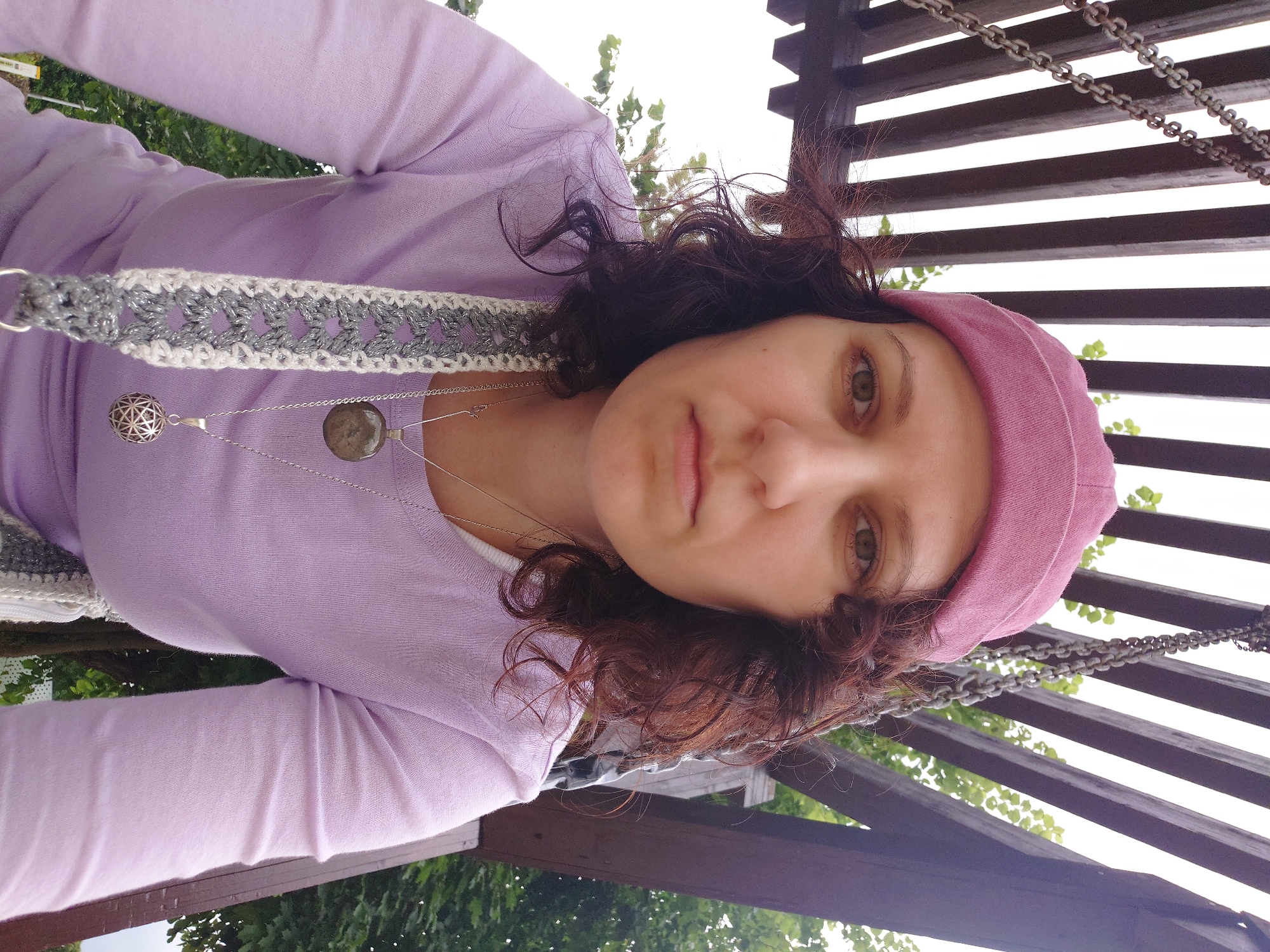 A medium shot shows a woman wearing a pink knit cap and a light purple long-sleeved shirt, looking directly at the camera. She has curly brown hair and is wearing two necklaces, one with a large, round, dark pendant and another with a smaller, ornate, silver pendant. A grey and white crocheted strap is visible across her chest. She appears to be sitting or standing on a swing set structure, indicated by the wooden beams and metal chains in the background, set against a backdrop of green foliage and a bright sky. The environment suggests an outdoor recreational area, possibly a park or a backyard. The lighting is diffused, indicating an overcast or shaded day. There is no discernible indication of the specific location within Kommunarka, Russia.