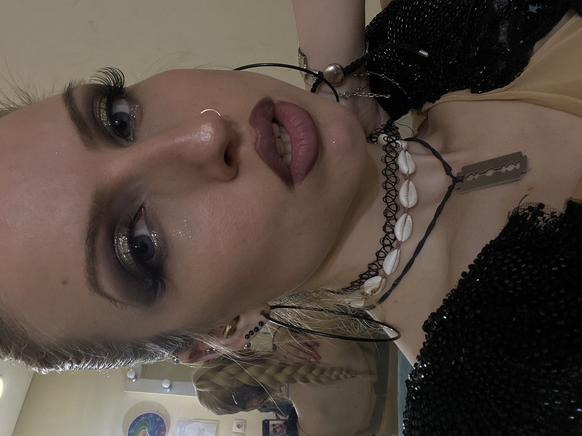 A close-up, slightly rotated photograph depicts a person with blonde hair styled in a braid, heavy makeup, and multiple piercings. The person has dark eye makeup with glitter accents, a nose ring, and multiple earrings. They are wearing several necklaces, including a choker with white shells and a necklace with a razor blade pendant. The person is wearing a black sequined top. The background appears to be a dressing room or backstage area, possibly in the city of Plovdiv, Bulgaria. A mirror reflects a partial view of the person and the surrounding area. Other details include jewelry on the person's wrist and visible parts of their clothing.