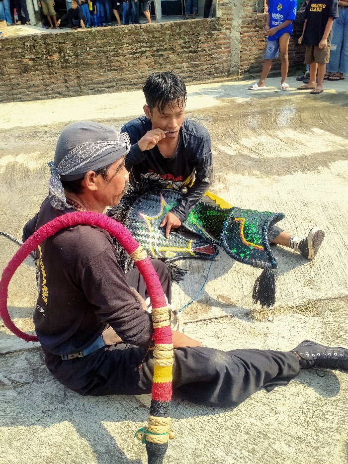 This outdoor scene in Sepat, Indonesia, depicts two males resting on a concrete surface adjacent to a weathered brick wall. The male on the left, an older individual wearing a patterned bandana, holds a large, red fabric-wrapped hoop with sections of yellow, black, and white cord. The male on the right, younger and with damp hair, is situated beside a decorated woven horse effigy, characteristic of Kuda Lumping, featuring green, black, and yellow designs. The younger male has an object at his mouth. Several other individuals are visible in the background against the wall. The ground exhibits damp patches, and strong sunlight casts distinct shadows.
