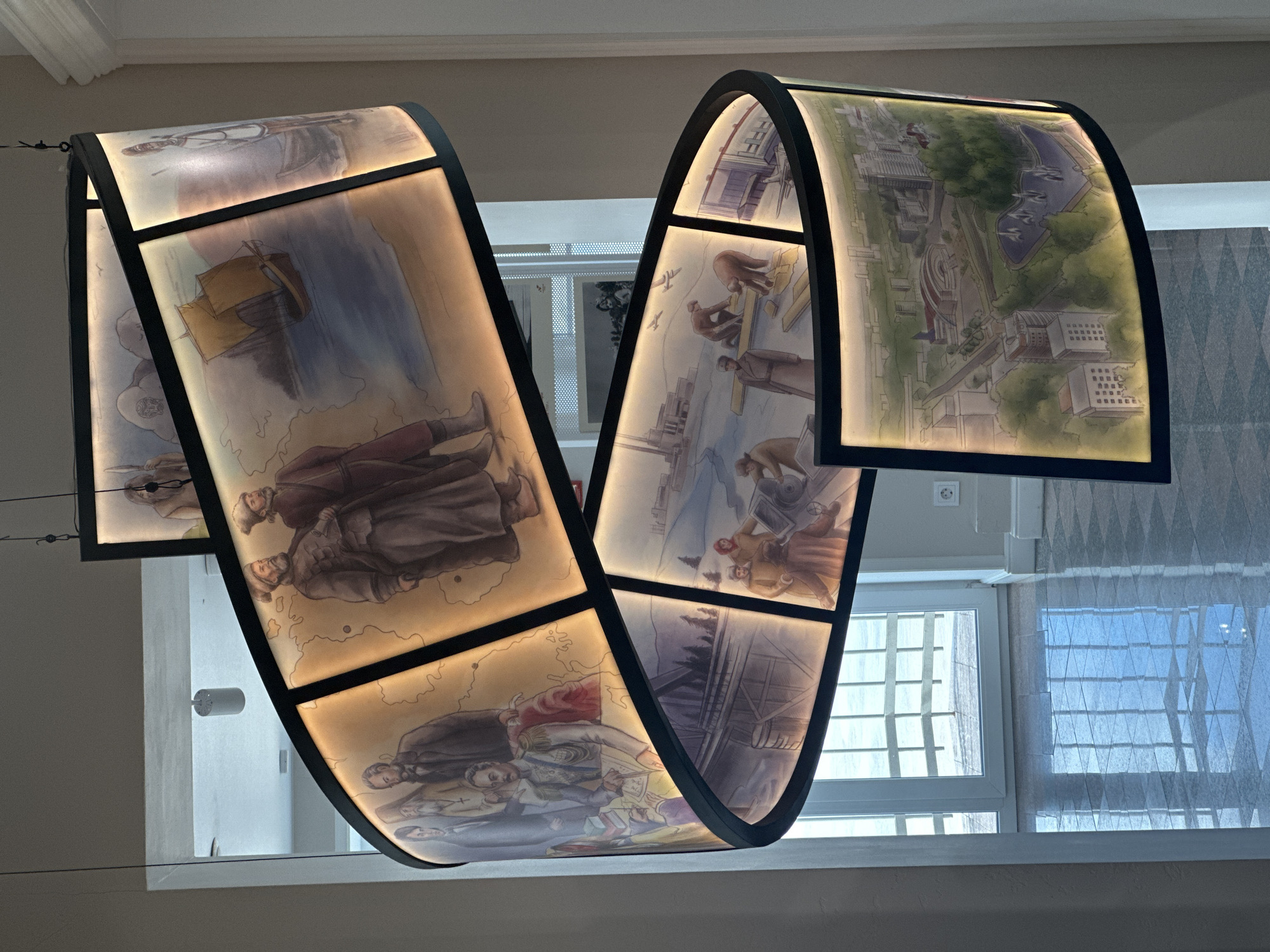 A spiral-shaped, illuminated art installation hangs from the ceiling in a well-lit indoor space. The installation features multiple rectangular panels depicting historical scenes. On the panels, one can see figures dressed in historical attire, a sailing ship on the water, industrial scenes with factories and workers, and a birds-eye view of a city with modern architecture. The installation appears to be in the Khabarovsk Regional Museum of Local Lore, named after N.I. Grodekov. The museum is located in Khabarovsk, Russia. The floor is tiled and highly reflective, showing a grid pattern from the windows. The background includes doorways and architectural elements of the building.