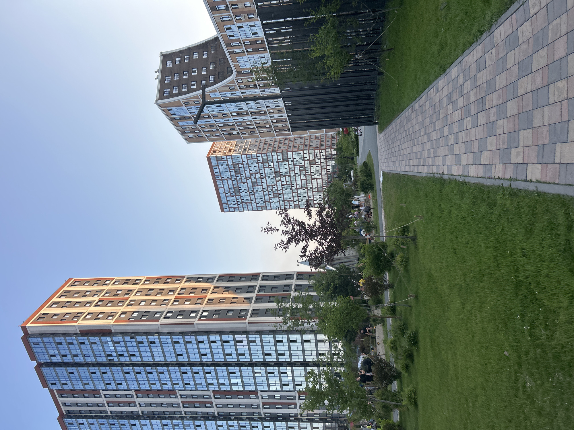 The image shows a modern residential complex with several high-rise buildings. The buildings have a mix of brick and glass facades, with balconies visible on some. A paved walkway runs alongside a manicured lawn, and several trees are planted in the area. In the background, people can be seen on the walkway and in the grassy area, some appear to be sitting on benches. The scene is outdoors, suggesting a pleasant day with clear skies, likely late afternoon or early evening given the lighting. The overall atmosphere appears to be one of urban living with integrated green spaces, a common feature in contemporary developments. The buildings are tall and sleek, indicative of a modern architectural style. There are no discernible text elements in the image.