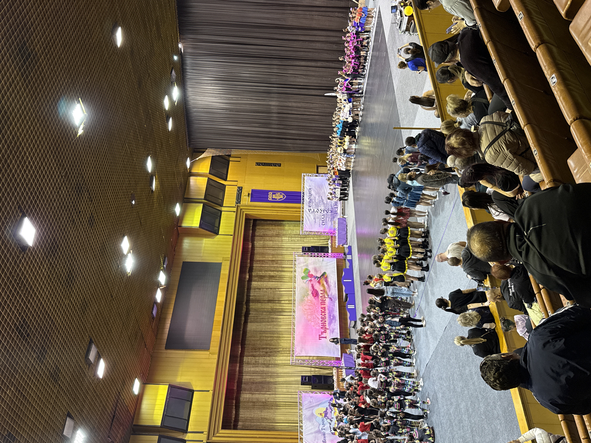 The image captures a dance competition venue in Unknown, Unknown. The scene features rows of young dancers lined up on the stage floor, facing the audience. They are diverse in their attire, with some wearing colorful performance outfits and others in more casual clothes. A stage is visible behind the dancers, adorned with banners that are written in an unreadable script. The hall appears to be full, with the audience seated in tiered rows of chairs, observing the dancers. The venue is lit by overhead lights, casting reflections on the floor.