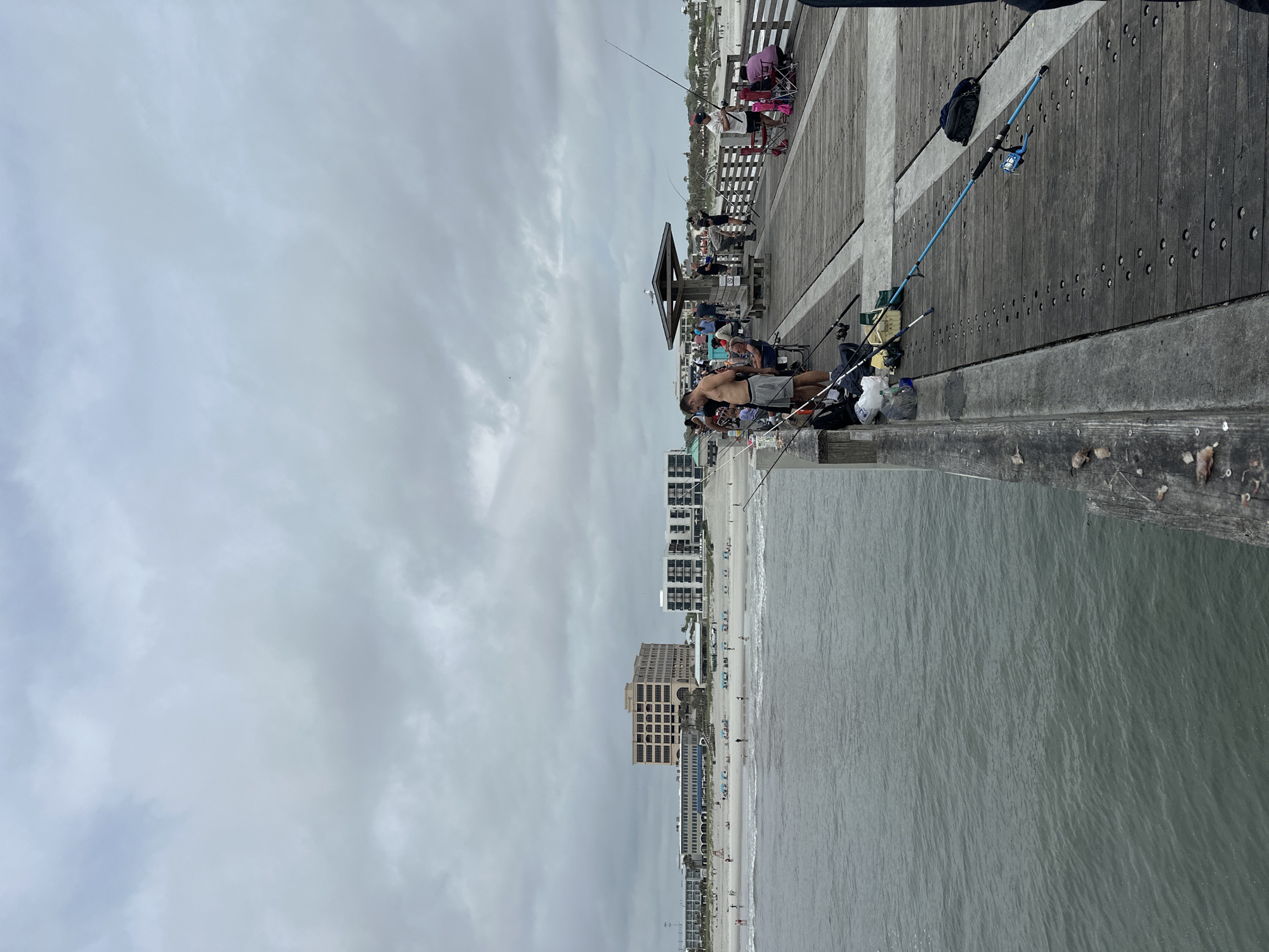 The photograph shows a scene at the Jacksonville Beach Fishing Pier in Jacksonville Beach, Florida, United States of America. The pier extends from the beach out into the ocean. The image is taken from the pier, looking towards the beach and cityscape. A group of people are fishing from the pier. The beach is crowded with people, and several tall buildings are visible along the shoreline. The sky is overcast with gray clouds. The pier itself is wooden with a gray concrete railing. Fishing rods and equipment are seen along the pier. A man is standing at the railing, apparently fishing.