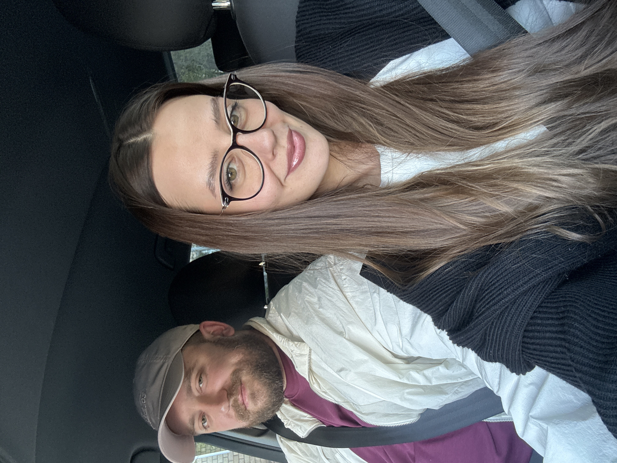 A woman with long brown hair and glasses sits in the passenger seat of a car, smiling towards the camera. She wears a white shirt under a black sweater. The car is likely a passenger vehicle, judging by the headrests. In the driver's seat, a man wears a light-colored baseball cap, a maroon t-shirt, and a light-colored jacket. The man is also smiling. Both occupants appear to be wearing seatbelts. The location in the city of Krasnoyarsk, Russia cannot be determined from this image alone.