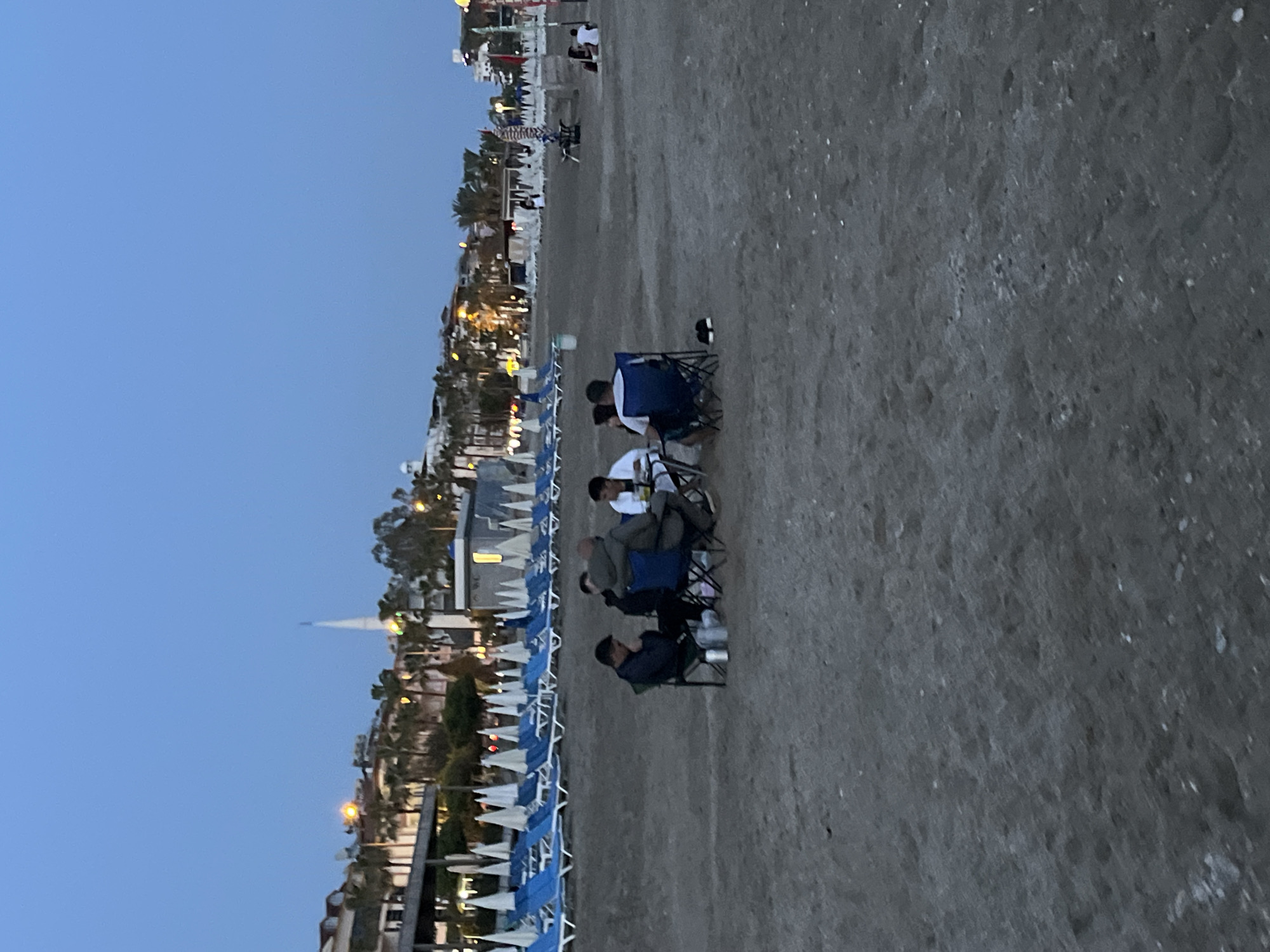 The image depicts a beach scene in Alanya, Turkey, at dusk. The sky is a clear, pale blue. A line of beach umbrellas and chairs, predominantly blue and white, stretches along the edge of the sand, adjacent to buildings, palm trees, and street lights. Four individuals are sitting in chairs further inland on the sand. One person wears a white shirt. The sandy beach occupies a significant portion of the foreground.