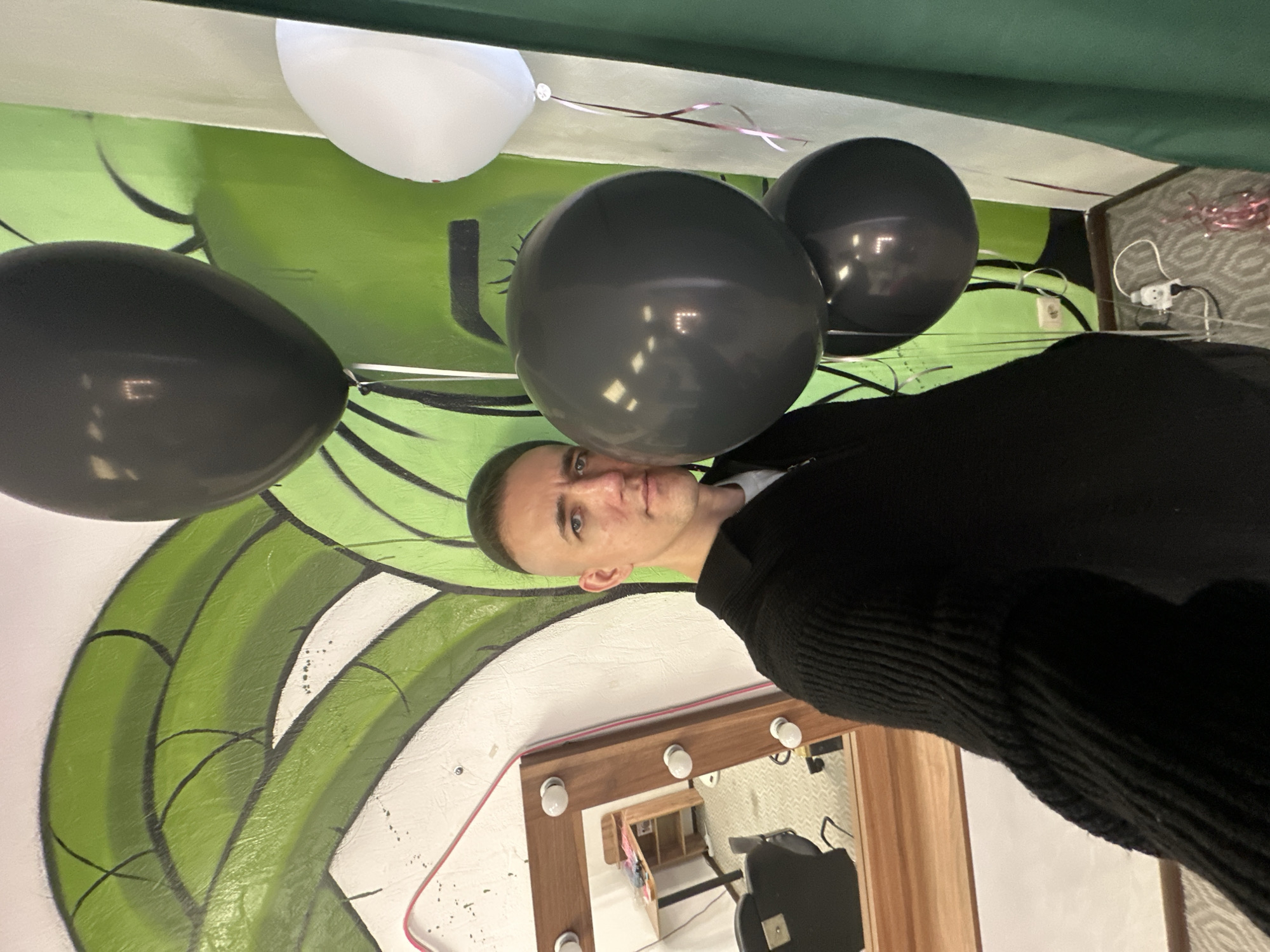 The scene depicts an interior space located in the city of Saratov, Russia. A single adult male is positioned centrally, facing the camera, consistent with a selfie perspective. He has a short, faded haircut with a green tint on the top section of his hair. He wears a black, ribbed, collared sweater with a zippered neckline over a lighter undergarment.
To the subject's left, a large, rectangular wooden-framed mirror is visible, featuring exposed light bulbs along its perimeter, indicative of a vanity or makeup station. The mirror's reflection shows a room containing a wooden desk or shelf, a black upholstered salon chair, and a patterned grey carpeted floor.
The background wall is adorned with a green and white mural featuring abstract, wavy, or leaf-like patterns outlined in black. Three black, spherical balloons are clustered to the subject's right and above, reflecting internal light sources. A portion of a white, spherical balloon with attached pink and dark red ribbons is visible on the far right edge of the frame, near the ceiling.
In the lower right corner, a white electrical wall outlet is present, with a white power strip and cable lying on the patterned grey floor. Scattered pink metallic tinsel or confetti is also visible on the floor in this area. A dark green curtain or fabric panel runs vertically along the right side of the frame.