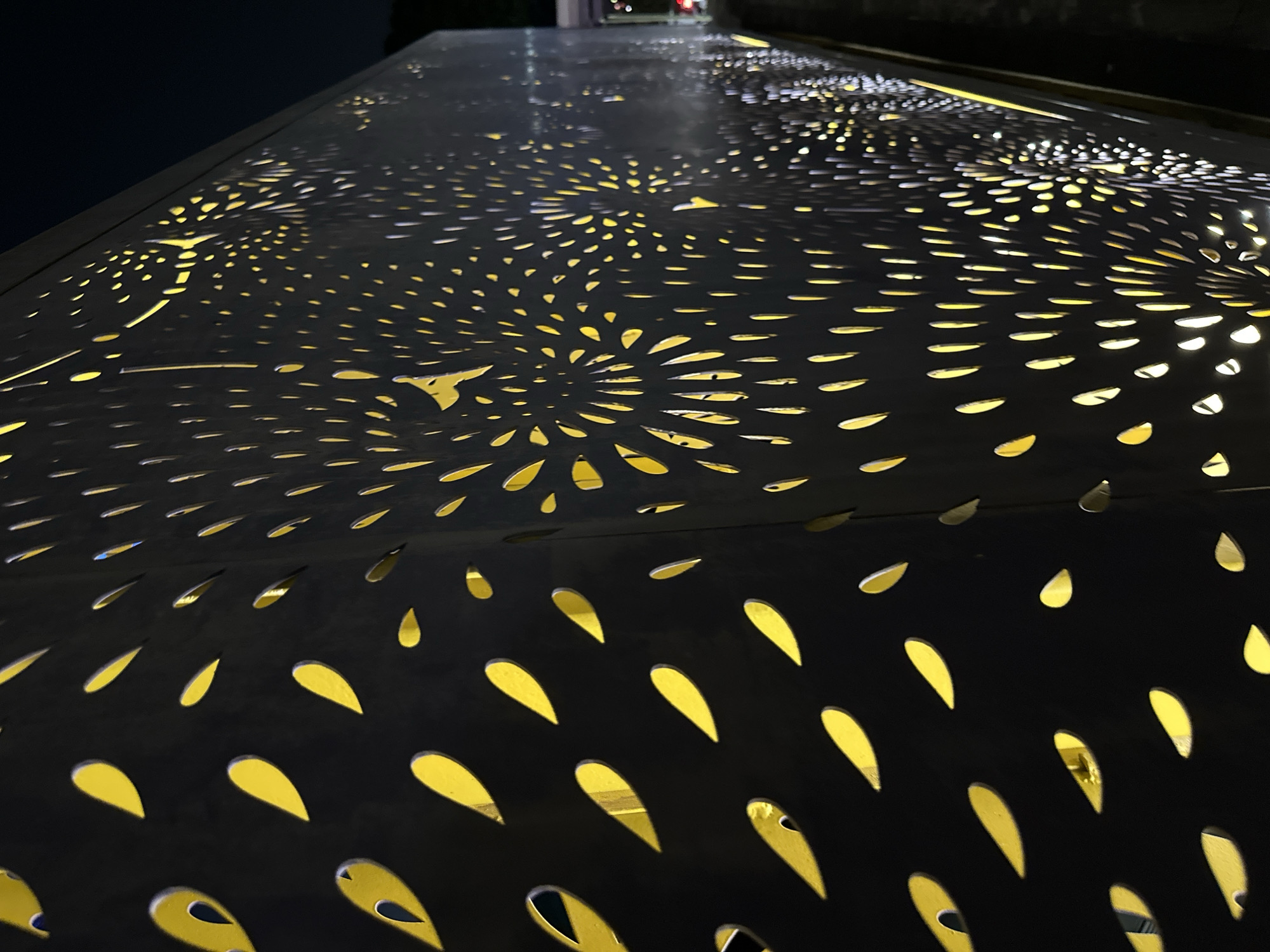 This is a close-up, eye-level, outdoor, night-time photograph of a dark metal facade featuring a pattern of cut-out shapes illuminated from behind with yellow light. The shapes on the left are elongated and teardrop-like, arranged in a staggered pattern. As the facade recedes into the distance on the right, the cut-outs transition into smaller, circular or oval shapes that form radial, almost floral or firework-like patterns. The lighting creates a dramatic effect, highlighting the perforations against the dark metal. The background is dark, suggesting it is nighttime, with a hint of a road or pathway visible on the far right edge. No people or specific landmarks are identifiable to pinpoint a precise location within Singapore.