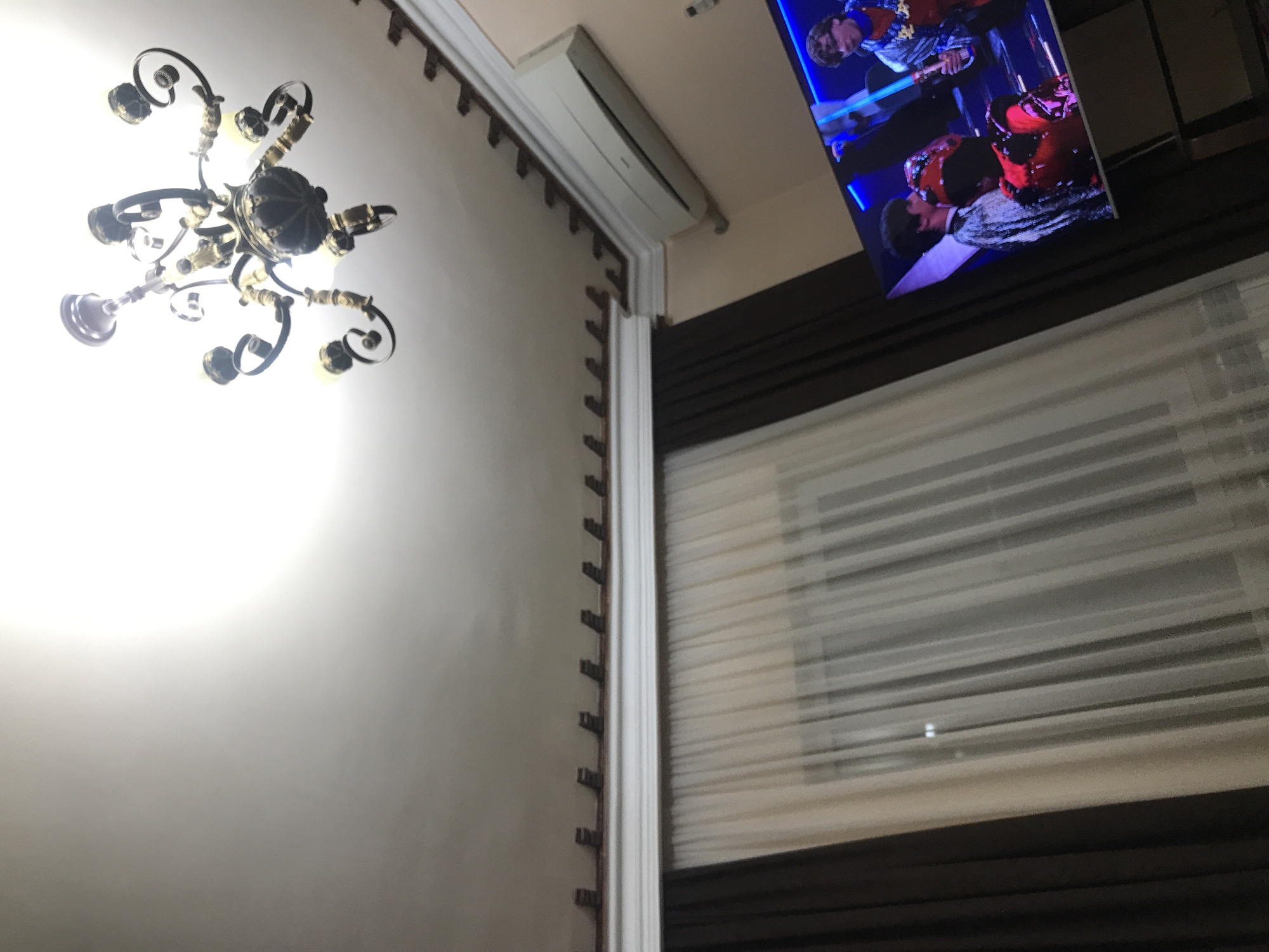 A low-angle shot depicts the interior of a room, likely in Yerevan, Armenia, as indicated by the user's request. A decorative chandelier hangs from the ceiling on the left. A wall extends from the left, adorned with molding. An air conditioning unit is mounted high on the wall near the ceiling. To the right, a television displays a music-related scene featuring people, possibly performers or a band. A window with horizontal blinds is visible below the television, with some light filtering through. The lighting suggests it could be either daytime or evening.