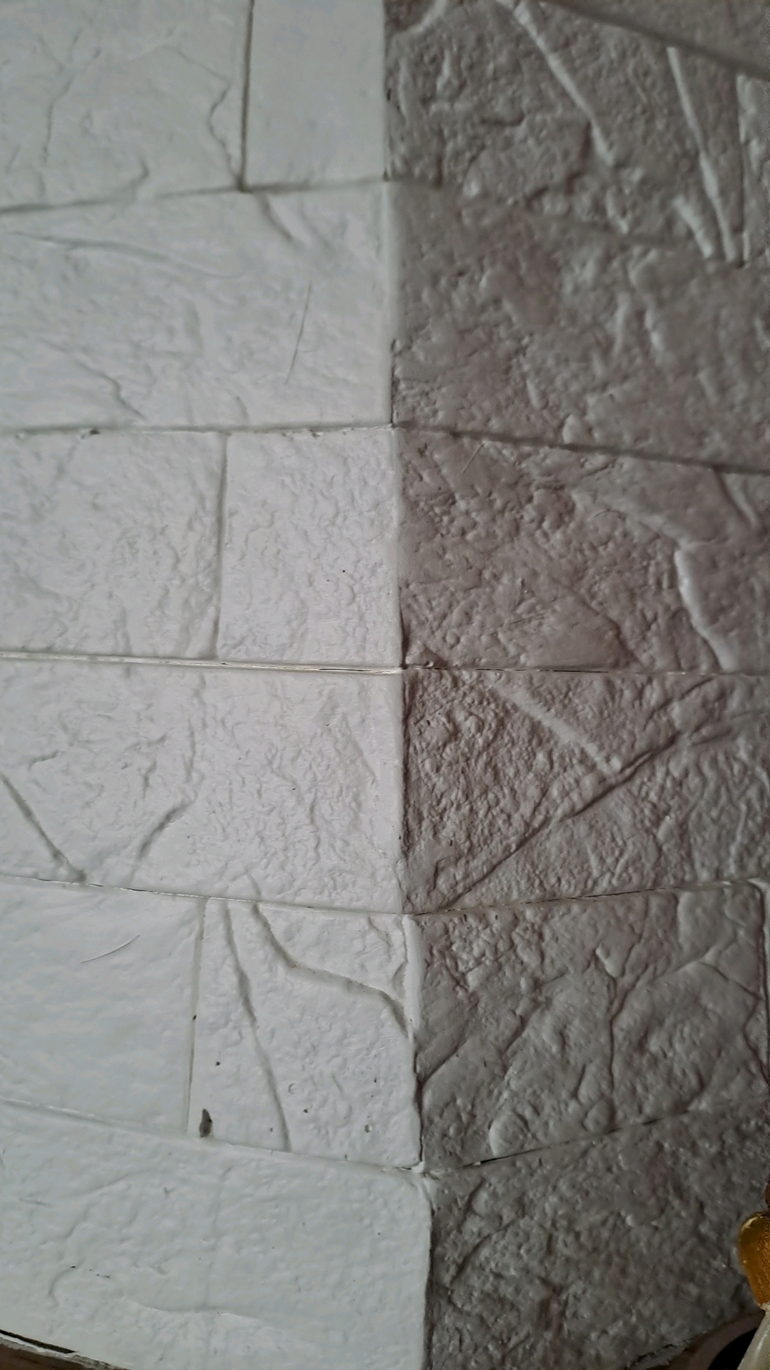The image shows a close-up, vertical view of a wall covered in textured faux brick panels. The wall is divided down the middle into two distinct color sections. The left side is a lighter shade, appearing off-white or pale gray, while the right side is a darker shade of gray, with visible shadows highlighting the texture. The faux bricks are irregular in shape and have a rough, stone-like surface with deep crevices and ridges. Horizontal and vertical grout lines are visible between the individual brick panels. The lighting appears to be coming from the top, casting shadows that emphasize the relief of the brickwork. There are no people or overt signs of activity in the image. The image does not provide any specific details that would indicate its location within the city of Kolomyia, Ukraine.