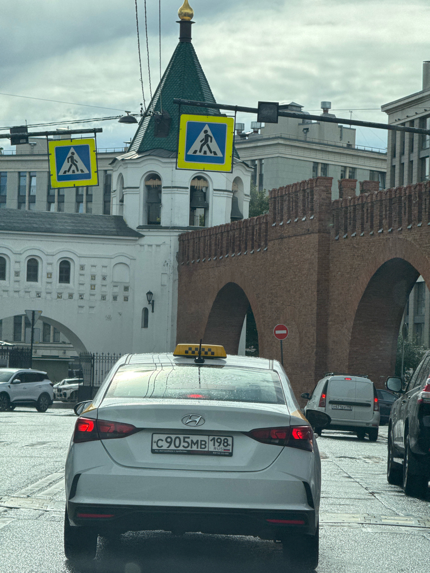 The image shows a view from a car on a city street in Saint Petersburg, Russia, with a focus on the rear of a white taxi. The taxi has a yellow illuminated sign on its roof with a checkerboard pattern and a Russian license plate displaying "C 905 MB 198". The car is on a wet road, indicating recent rain.
In the background, there is a distinctive white building with arched windows and a green-domed tower topped with a golden spire. Adjacent to this building is a red brick structure with large arches, resembling a fortress wall or an old bridge. Two pedestrian crossing signs, displaying a silhouette of a person walking within a blue square and a yellow border, are visible suspended over the road. Further in the background, other vehicles and buildings are present, including a white van with a license plate that appears to read "Y471EY 198" and other cars. The sky is overcast.