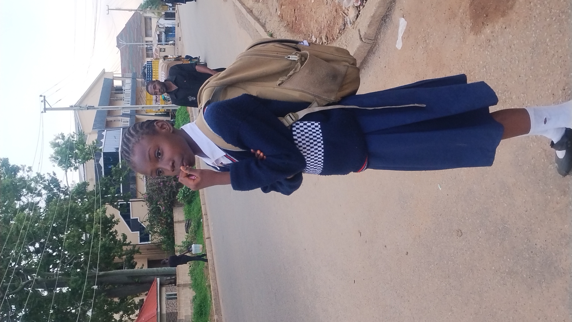 The primary subject of the image is a young girl, likely a student, standing on the side of a road in Jos, Nigeria. She is wearing a school uniform that includes a white collared shirt, a dark blue V-neck sweater with a prominent white and dark blue checkered pattern around the waist, and a dark blue pleated skirt. She also wears white socks and black Mary Jane-style shoes. A light brown backpack is slung over her shoulders. Her hair is styled in neat braids, and she looks directly at the camera with a neutral expression, her right hand near her mouth.
Behind the girl, slightly to her right, a man in a dark short-sleeved shirt is visible, also looking towards the camera. Further in the background, another person can be partially seen. The scene is set on an urban or semi-urban street. The ground features a visible asphalt road alongside a lighter-colored dirt or gravel shoulder. Buildings, some multi-storied with visible windows and balconies, line the street. Lush green trees and other foliage are present, along with utility poles supporting overhead power lines. The sky is bright, indicating it is daytime, possibly with an overcast or hazy atmosphere that provides soft, diffused lighting. No discernible text is visible within the image.