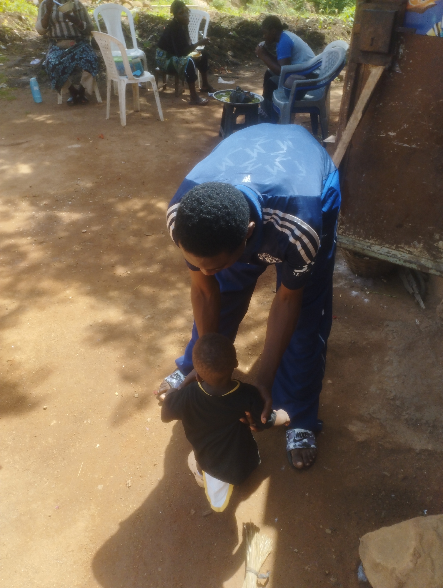 In a dusty, outdoor setting in Jos, Nigeria, a man wearing a blue tracksuit with white stripes down the sleeves and sides, along with black Nike slides, is bent over a small child. The child, dressed in a black t-shirt and white shorts with yellow trim, is facing away from the camera. The man appears to be assisting the child, possibly helping them to stand or walk. The ground is composed of reddish-brown dirt with scattered pebbles and fallen leaves, and dappled sunlight creates shadows on the ground. In the background, several white plastic chairs are visible, and further back, people are seated, suggesting a gathering or meal. One person in the background is wearing a blue shirt. To the right of the scene, a large, rustic, metal structure is present, possibly a shed or equipment. A bundle of straw or dry grass lies on the ground near the child's feet.
