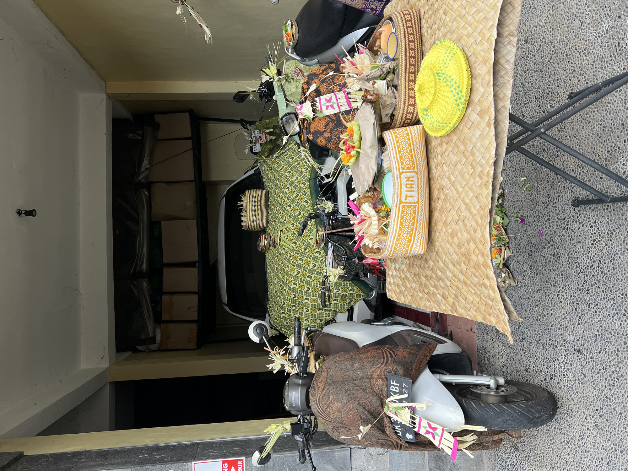 The image captures a street scene in the Ubud District of Indonesia. A white and brown Honda scooter is positioned in front of a building's opening. Both the scooter and a nearby folding table are adorned with offerings in woven baskets and fabrics, which are traditional Balinese decorations. The table is covered in a large, woven mat. The scooter's license plate is visible. The building's interior is dimly lit, with boxes visible on a shelf in the background. The street surface is paved with small stones.