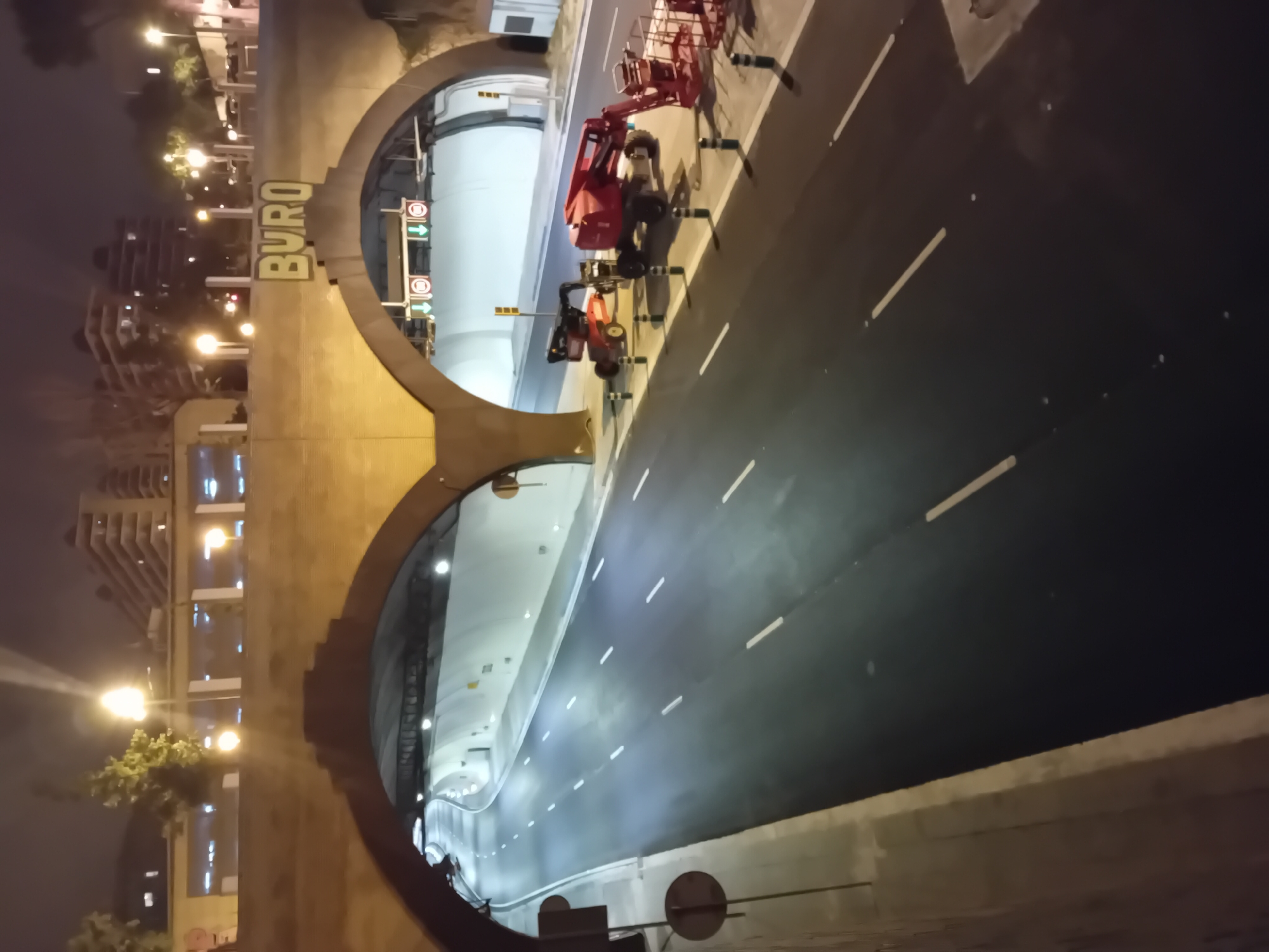 Captured from a high vantage point at night, the image shows the Buro Tunnels in Barcelona, Spain. The tunnel entrances are framed by distinctive, curved architectural structures, with the name "BURO" emblazoned on one. Inside the brightly lit tunnels, the road surface is dark, marked with white lane lines. Construction or maintenance vehicles are parked near the entrance of the upper tunnel. The surrounding environment includes buildings with lit windows and streetlights, indicating an urban setting.