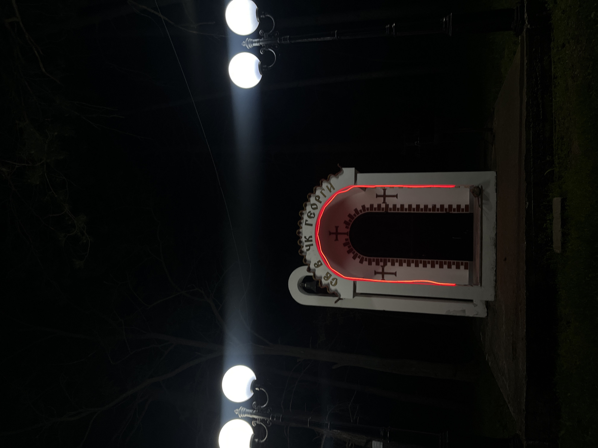 The image depicts a religious monument at night in Sungurlare, Bulgaria, under the illumination of two double-globe street lamps. The monument, outlined in red light, features Cyrillic script on its rounded top and crosses on its facade. It has a narrow, dark opening. The surrounding environment is dark with trees visible in the background.