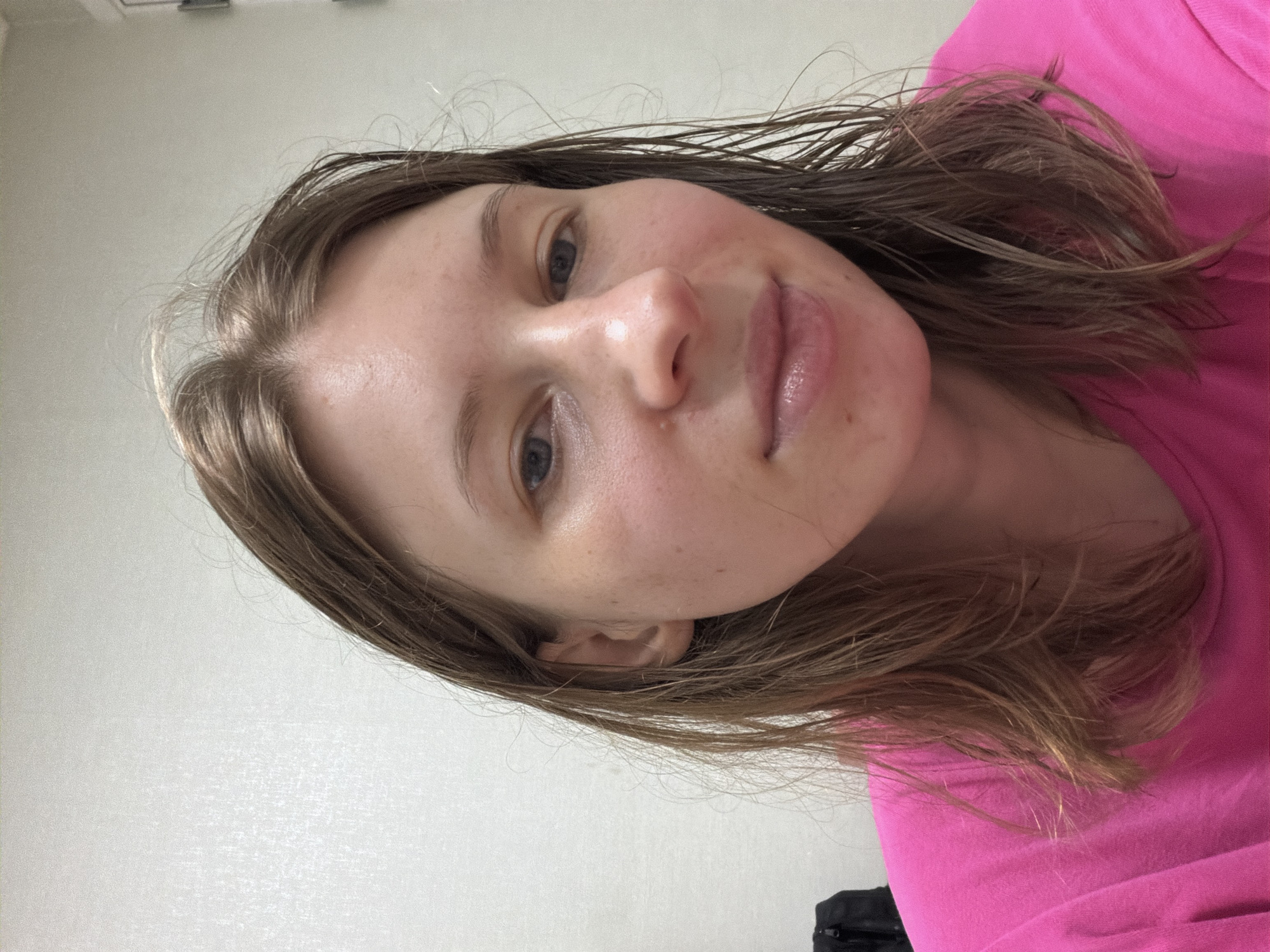 The image shows a close-up, head-and-shoulders shot of a young Caucasian woman with light brown, wet hair, wearing a bright pink t-shirt. She is looking directly at the camera with a neutral expression. Her skin appears fair with some natural freckles. The background is a plain, lightly textured off-white wall, likely indoors. To the far left of the frame, a black bag or backpack is partially visible. The lighting is soft and diffused, suggesting an indoor setting without direct sunlight. There is no indication of the city of Dongducheon-si, South Korea.