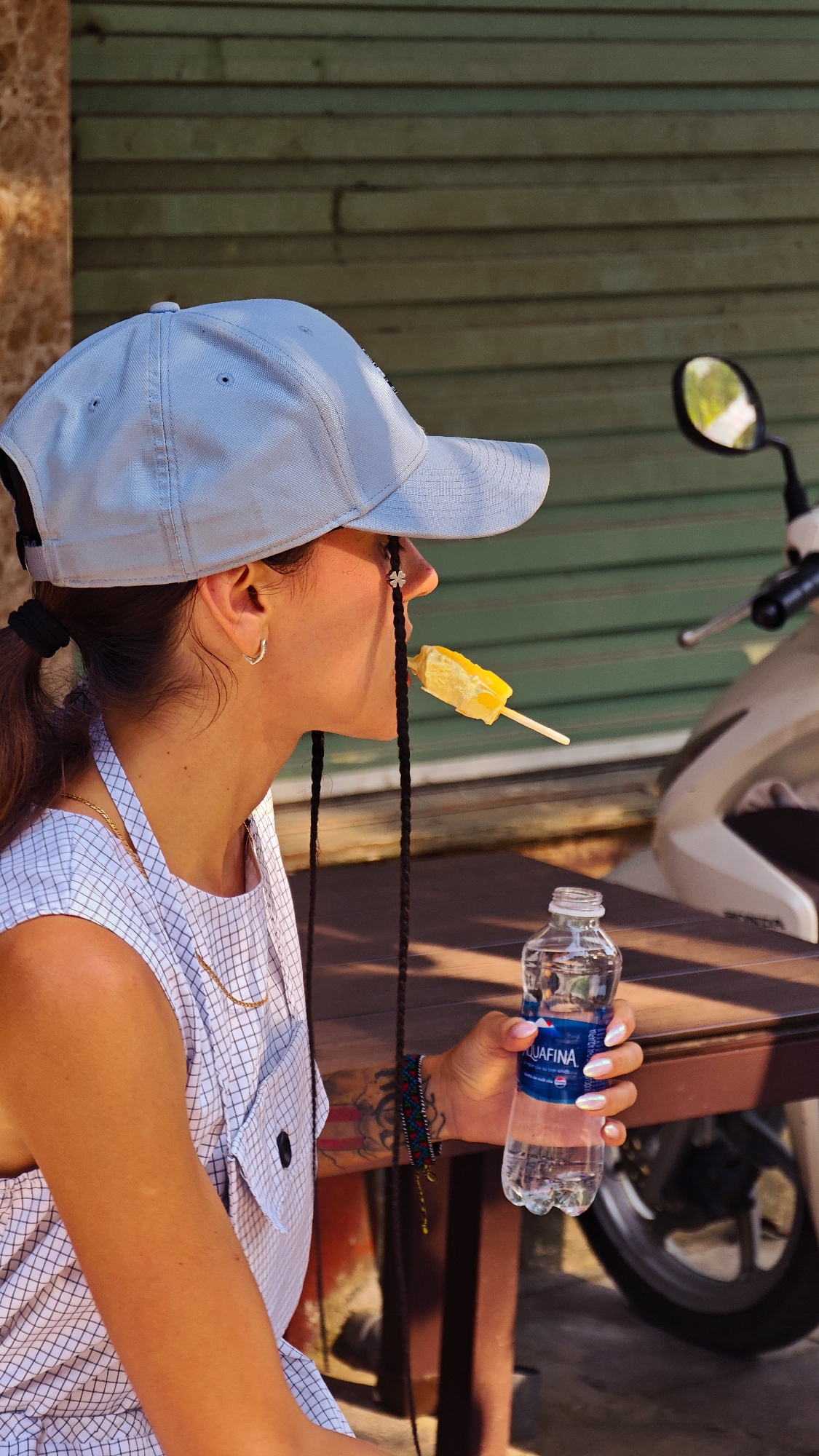In Bắc Hà, Vietnam, a young woman is seated at a table outdoors. She wears a light blue baseball cap pulled low, a white checkered sleeveless shirt, and has long dark braids extending from under her cap. She is holding a water bottle with the label "AQUAFINA" in her right hand and eating a yellow popsicle. A motorcycle is parked behind the table. The backdrop appears to be a green wooden wall.