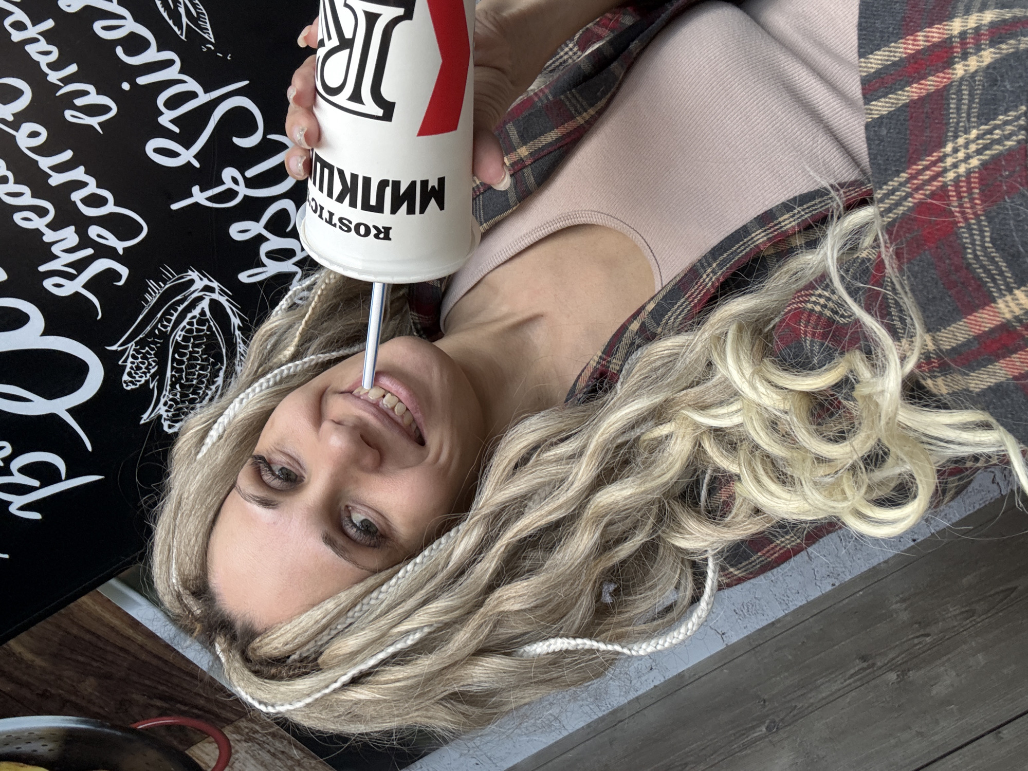A woman with blonde hair and white braids is pictured indoors, holding a "ROSTIC'S" cup with a straw. She is wearing a light pink top and a plaid shirt. Next to her is a black surface with white writing and a corn drawing. In the background, a metal pan with food is visible, along with a wooden floor. The city of Rusanovo, Russia is not explicitly identifiable in the image.