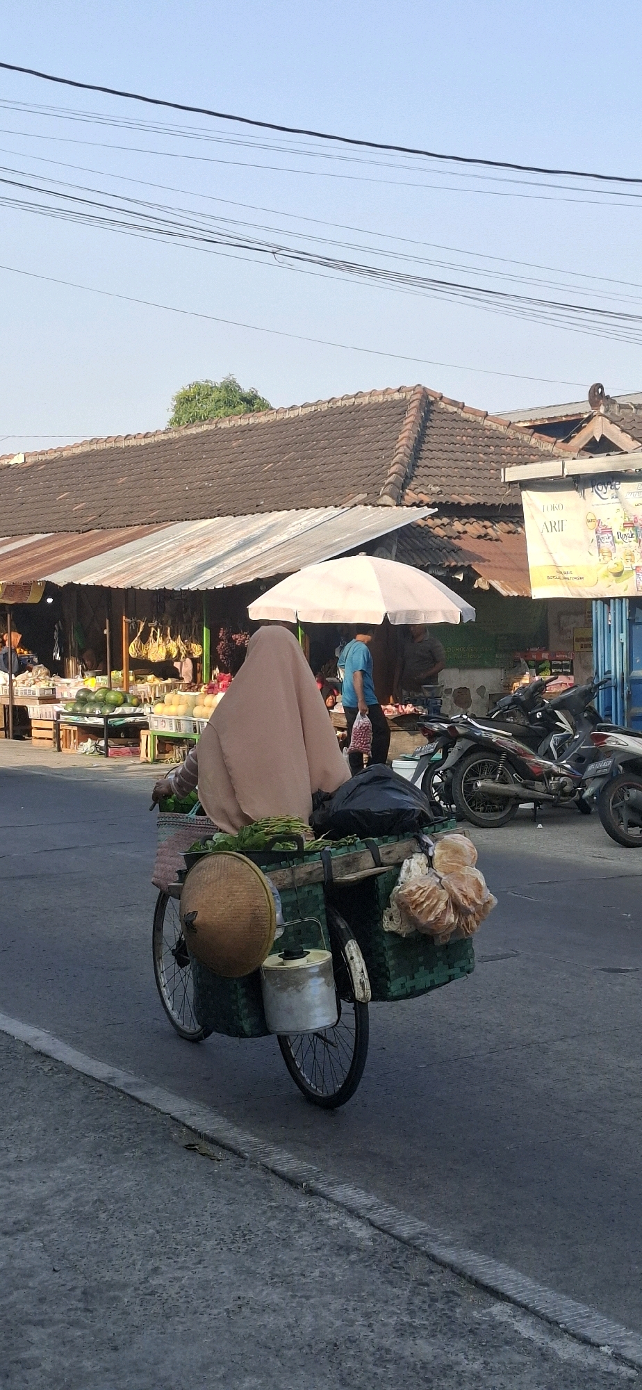 This is a full-shot, eye-level photograph taken on a sunny day in Desa Dibal, Indonesia, showing a person on a bicycle loaded with goods. The subject, wearing a light brown hijab and a pinkish-brown robe, is positioned in the center of the frame, facing away from the camera and riding the bicycle. The bicycle has two large green woven baskets attached to the rear, one on each side, carrying vegetables and other items. A conical straw hat is hanging on the left basket, and a metal container is attached below it. A black plastic bag and some light-colored bundles, possibly food items, are also visible in the baskets.
The background reveals a market scene. Several stalls with tiled roofs and tin awnings are visible, displaying various goods including fruits and vegetables like watermelons and what appear to be melons or gourds. Two motorcycles are parked on the right side of the frame. In the distance, a person in a blue shirt is visible walking. The environment appears to be an outdoor market or roadside selling area, with a road in the foreground and a clear blue sky above with a few power lines. The image is focused on the person and their bicycle, with the market elements serving as a contextual backdrop.