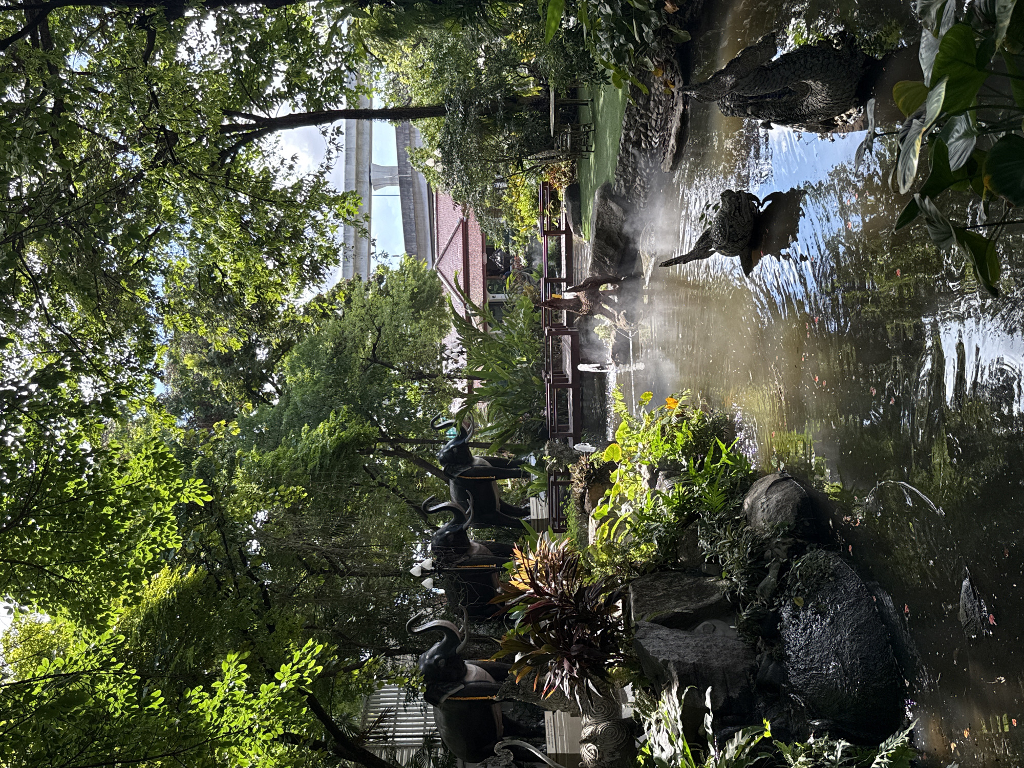 The scene depicts a garden in Samrong Nuea Subdistrict Municipality, Thailand, with a pond as a central feature. The pond contains sculptures of birds and what appears to be warm water, suggesting hot springs. Trees surround the area, creating a lush environment with natural rocks and plants. A bridge crosses the pond, and a traditional Thai-style house is visible in the background. In the distance, an elevated highway is seen above the treeline. There are decorative elephant sculptures near the ground level.