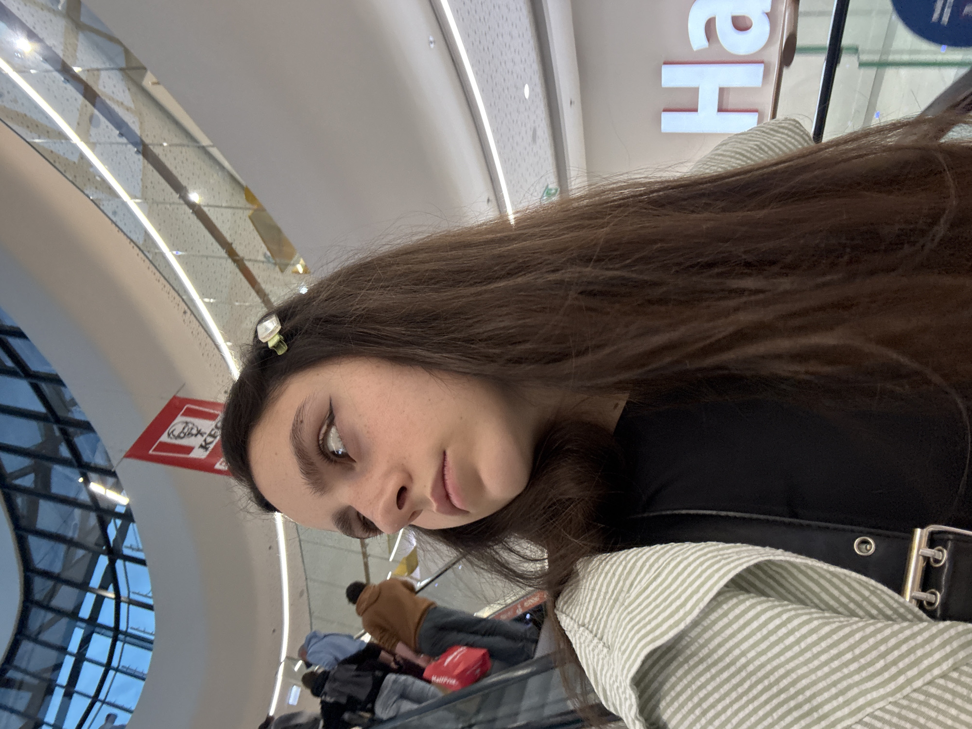 The image shows a young woman with long brown hair, adorned with a small white clip, looking to her right. She is wearing a black top and a light green and white striped cardigan. A black bag with silver hardware is visible across her shoulder. The background suggests an indoor setting, likely a shopping mall. People are seen on an escalator, some carrying bags. A red KFC banner hangs in the upper part of the frame, and a large sign with the letters "Ha" in white is visible further in the background, indicating a commercial establishment. The architecture includes a modern, multi-level design with a glass ceiling structure. The lighting appears to be artificial, typical of an indoor commercial space. The overall impression is that of a casual moment captured within a bustling shopping environment, potentially in Opole, Poland, given the presence of a prominent retail establishment that might be found in such a city.