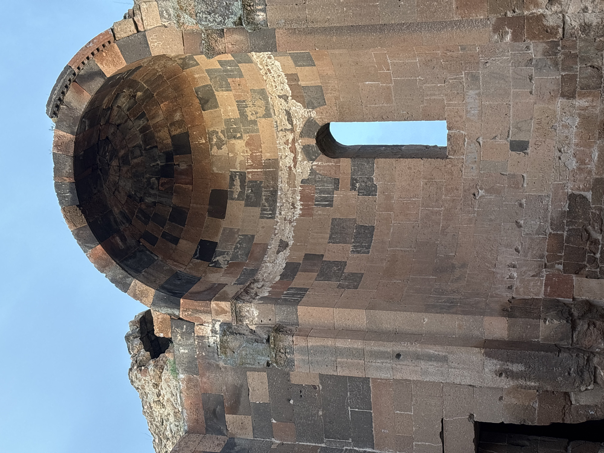The image shows a detail of the ruins of a medieval Armenian church, specifically the Church of St. Gregory of Abughamrents in Ani, a ruined medieval Armenian city now located in present-day Turkey. The photograph is taken from a low angle, looking upwards at the structure.
The focal point is the cylindrical dome of the church, constructed from alternating light and dark-colored stone blocks. The dome's exterior features a row of decorative elements. The interior of the dome is visible, showing the concentric rings of stone that form its structure.
To the right of the dome, a section of the church wall remains, also constructed from light and dark stone. A rectangular window is visible in the wall.
The sky is visible through the window and partially surrounding the structure, indicating the building is in ruins. The overall impression is one of aged construction.