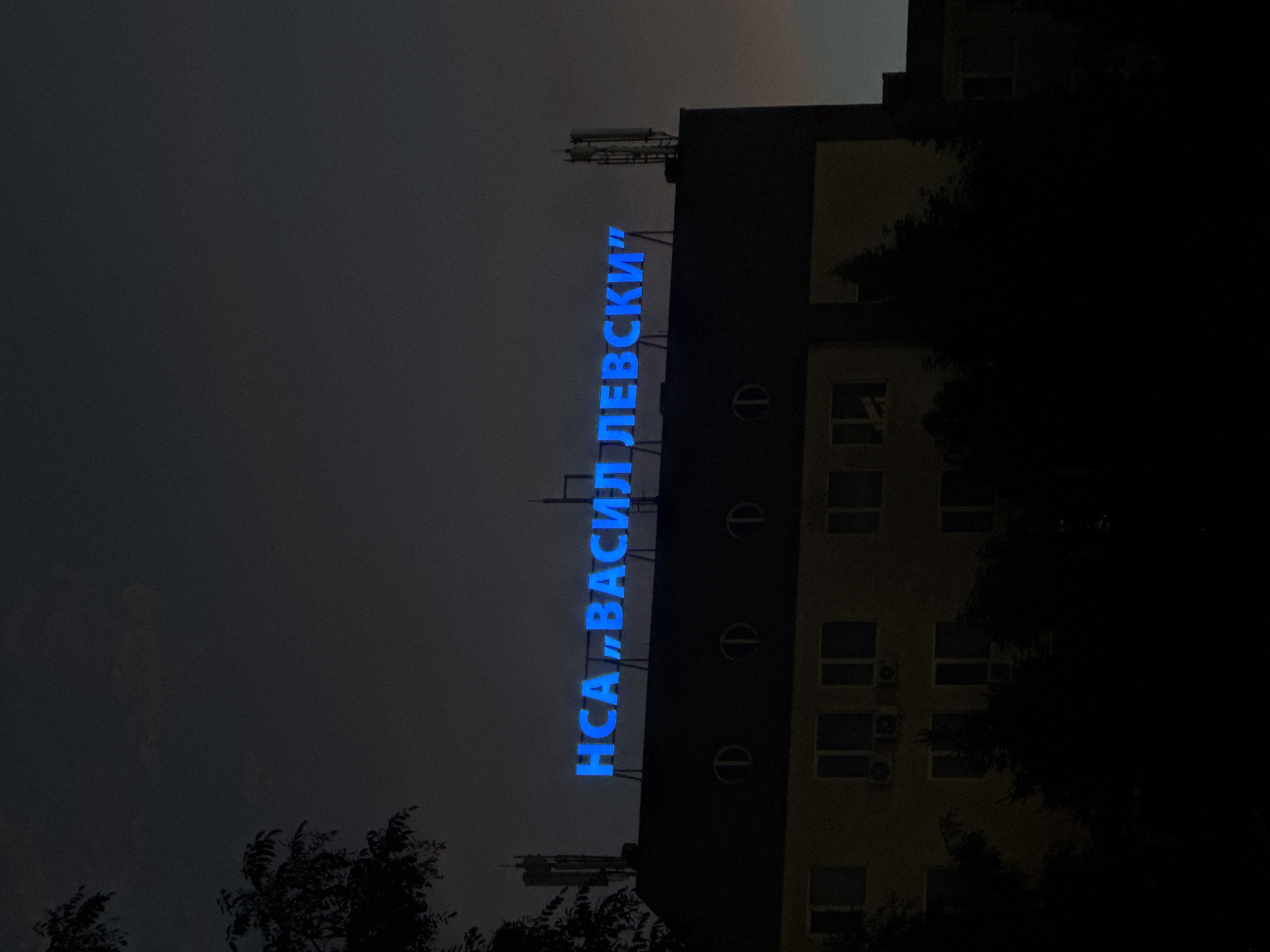 The image shows a building with a neon sign that reads "НСА „ВАСИЛ ЛЕВСКИ”" in blue letters. The building appears to be at dusk or night, as the sky is dark and the sign is illuminated. There are also antennas on the roof of the building, and trees in the foreground, silhouetted against the building. The building has several windows and what appear to be air conditioning units below some of them. Based on the signage, the building is likely the National Sports Academy "Vasil Levski" in Sofia, Bulgaria.