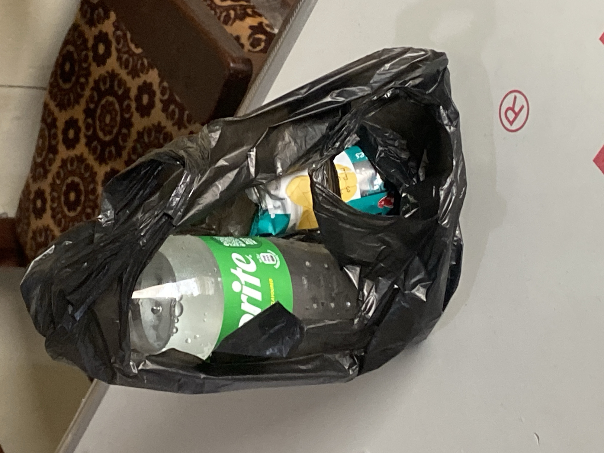 A black plastic bag rests on a white surface. Inside the bag, a clear plastic bottle of Sprite is visible, partially obscured by the bag. Beside the bottle, several small, foil-wrapped packets are present. In the background, a patterned sofa or chair can be seen. A red symbol resembling a circled 'R' is imprinted on the white surface below the bag. The image appears to be taken indoors, potentially in Jauro Yinu, Nigeria, given the context provided.