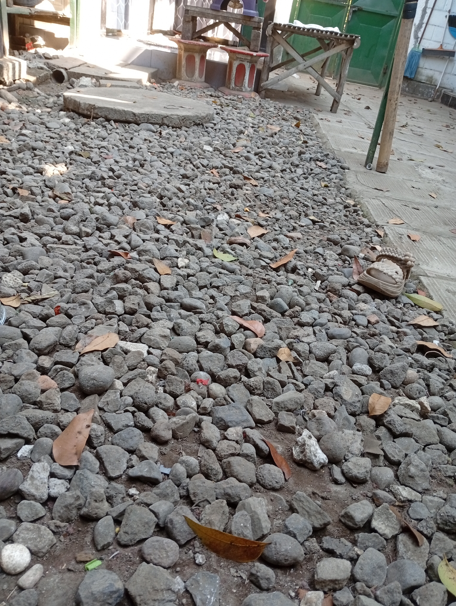 The image shows a ground-level view of an outdoor area covered in gray gravel. In the background, there is a concrete manhole cover and a small pipe outlet. To the right, there is a concrete path and a wooden structure with a green door. A few scattered dry leaves are visible on the gravel and concrete. A worn-out shoe is partially visible on the gravel. The scene appears to be in a backyard or yard area in Balung, Indonesia, characterized by its utilitarian and somewhat unkempt appearance.
