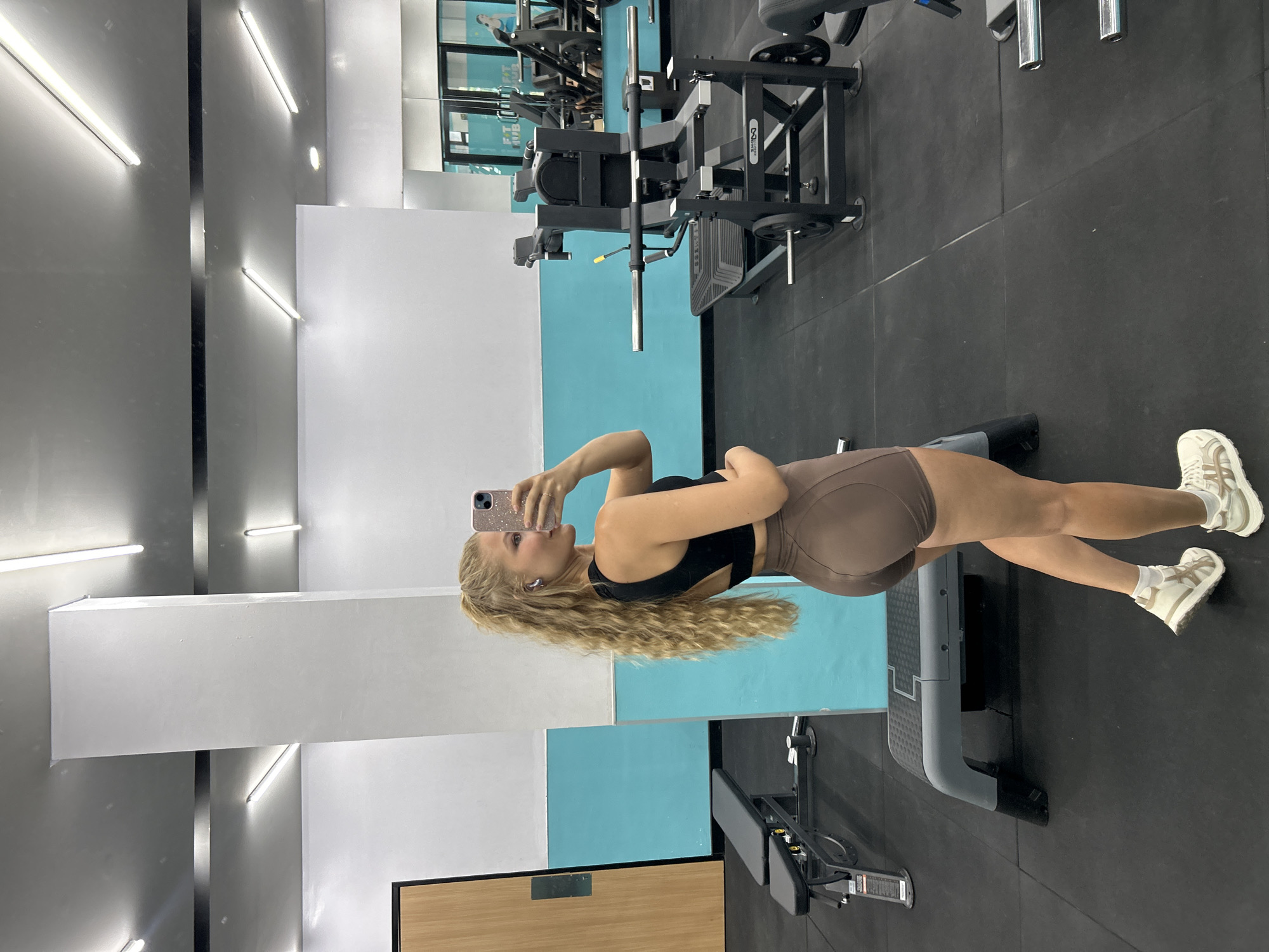 This media file is a rotated indoor photograph of a young woman taking a selfie in a gym. The image is rotated 90 degrees counter-clockwise, with the woman standing horizontally on the right side of the frame, facing left.
The main subject is a woman with long, wavy blonde hair, wearing a black sports bra and light brown/tan form-fitting cycling shorts. She holds a light-colored smartphone with a sparkling case in her left hand, angled towards a mirror to capture her reflection. Her right hand rests on her hip. She has small earrings and appears to be looking at her phone screen.
The setting is a modern gym. The floor is covered with dark grey or black rubber tiles. The walls feature sections of white, light blue, and grey/silver panels, many of which are reflective mirrors. Several bright, linear fluorescent lights are visible on the walls and ceiling, illuminating the space.
In the background, various pieces of black gym equipment are present. A grey and black weight bench is positioned on the floor near the woman's legs. Further back, a larger black weight machine, possibly a leg press or similar, is seen with silver accents and weight plates. The mirrors reflect more of the gym interior, including additional equipment and lights. Faintly visible text in the mirror's reflection includes the words "FIT HUB" on what appears to be a sign or logo. A section of a wooden panel or counter is visible in the bottom left corner of the frame.
The overall scene depicts a fitness enthusiast documenting her presence in a well-equipped gym. The location context provided is Kuta Selatan, Indonesia.