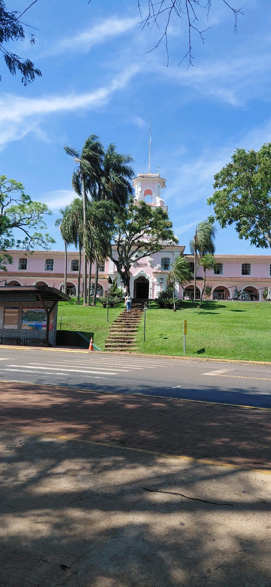 The image depicts the Hotel Cassino Tabajara, a pink historic hotel located in Foz do Iguaçu, Brazil. The hotel features a prominent tower with a spire and arched windows. In front of the hotel, a wide, manicured lawn leads up to a stone staircase. Two individuals are walking up the stairs towards the hotel entrance. To the left of the stairs, a bus stop shelter is visible, with a map displayed inside. The scene is set against a bright blue sky with scattered white clouds. Palm trees and other lush green vegetation surround the hotel. In the foreground, a paved road with a pedestrian crossing runs across the bottom of the frame, with shadows of trees cast upon it. A small orange traffic cone is present near the bus stop.