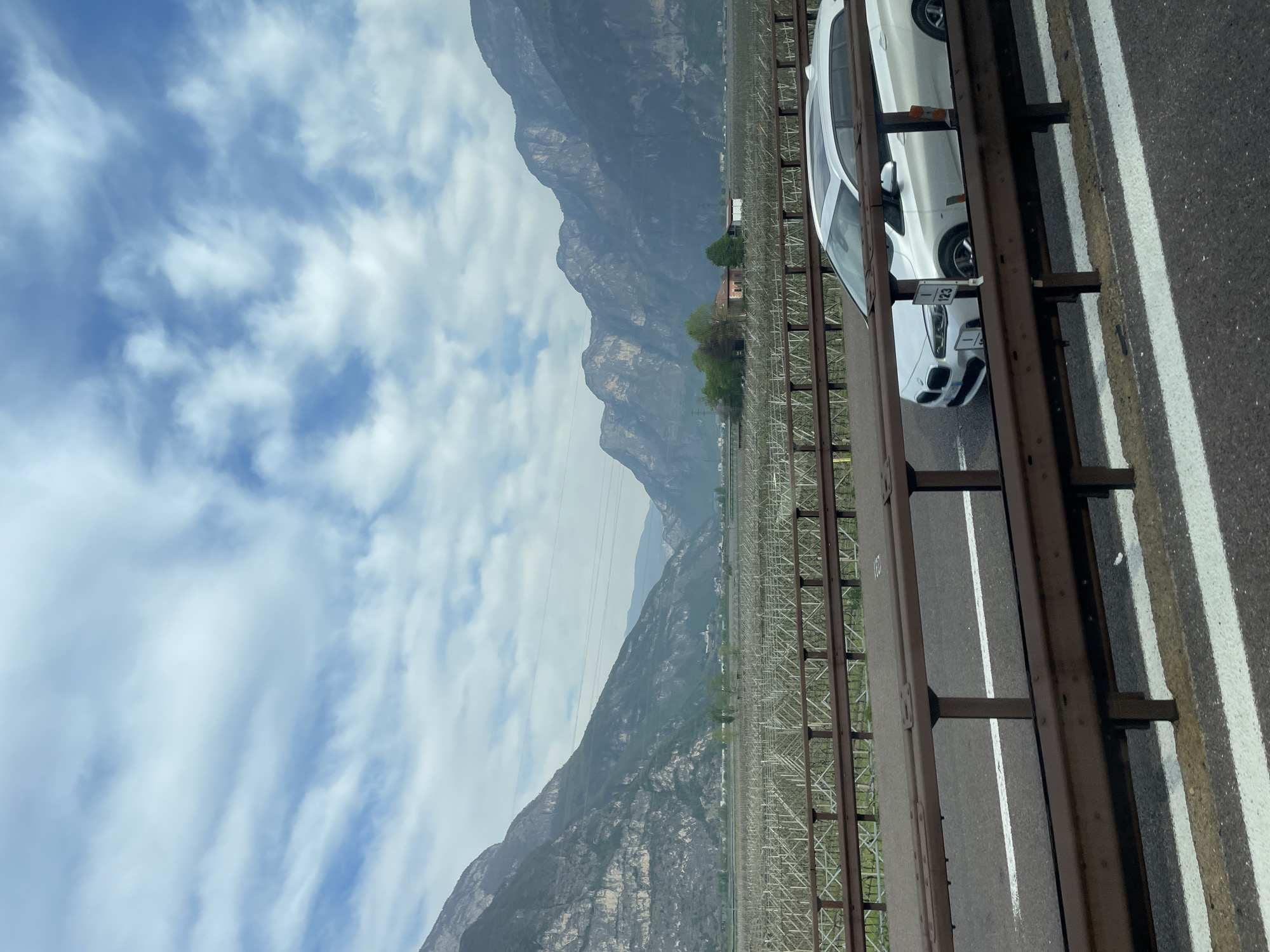 The image depicts a view from a bridge in Nave San Rocco, Italy. In the foreground, a white BMW car is seen parked on the bridge's shoulder, separated from the traffic lane by a metal barrier. The highway asphalt shows white lane markings. In the background, a mountainous landscape under a partly cloudy sky dominates the view. A large agricultural field with trellises is also visible, suggesting a vineyard or orchard. Power lines stretch across the scene.