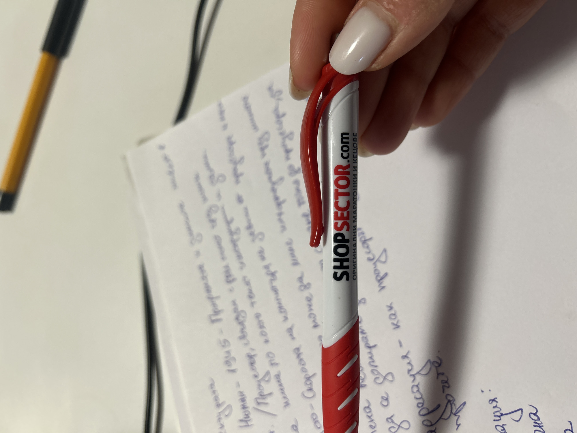 The image shows a hand holding a red and white pen with the logo "SHOPSECTOR.com ОРИГИНАЛНИ МАРАТОНКИ И КЕЦОВЕ." The pen is positioned over a piece of paper with handwritten notes in blue ink. A yellow and black pen is also visible in the background on a white surface. No specific details are available in the image that suggest the location in the city of Burgas, Bulgaria.