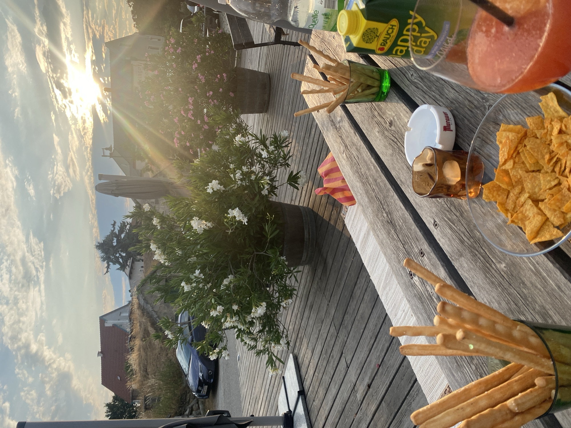 The image depicts a table setting on a wooden deck, with a warm, late afternoon sun casting long shadows and creating lens flare. In the foreground, a wooden table holds a glass of reddish-orange beverage with a straw, a bowl of tortilla chips, an ashtray, a small glass votive with a lit candle, a green glass filled with breadsticks, and a carton of "Rauch happy day" juice. Behind the table, two large potted plants, one with white flowers and the other with pink flowers, are visible. A blue car is parked on a gravel area in the background, with houses and trees situated on a gentle slope. The overall ambiance suggests a relaxed outdoor setting, possibly a restaurant or a private patio, in Purbach, Austria.